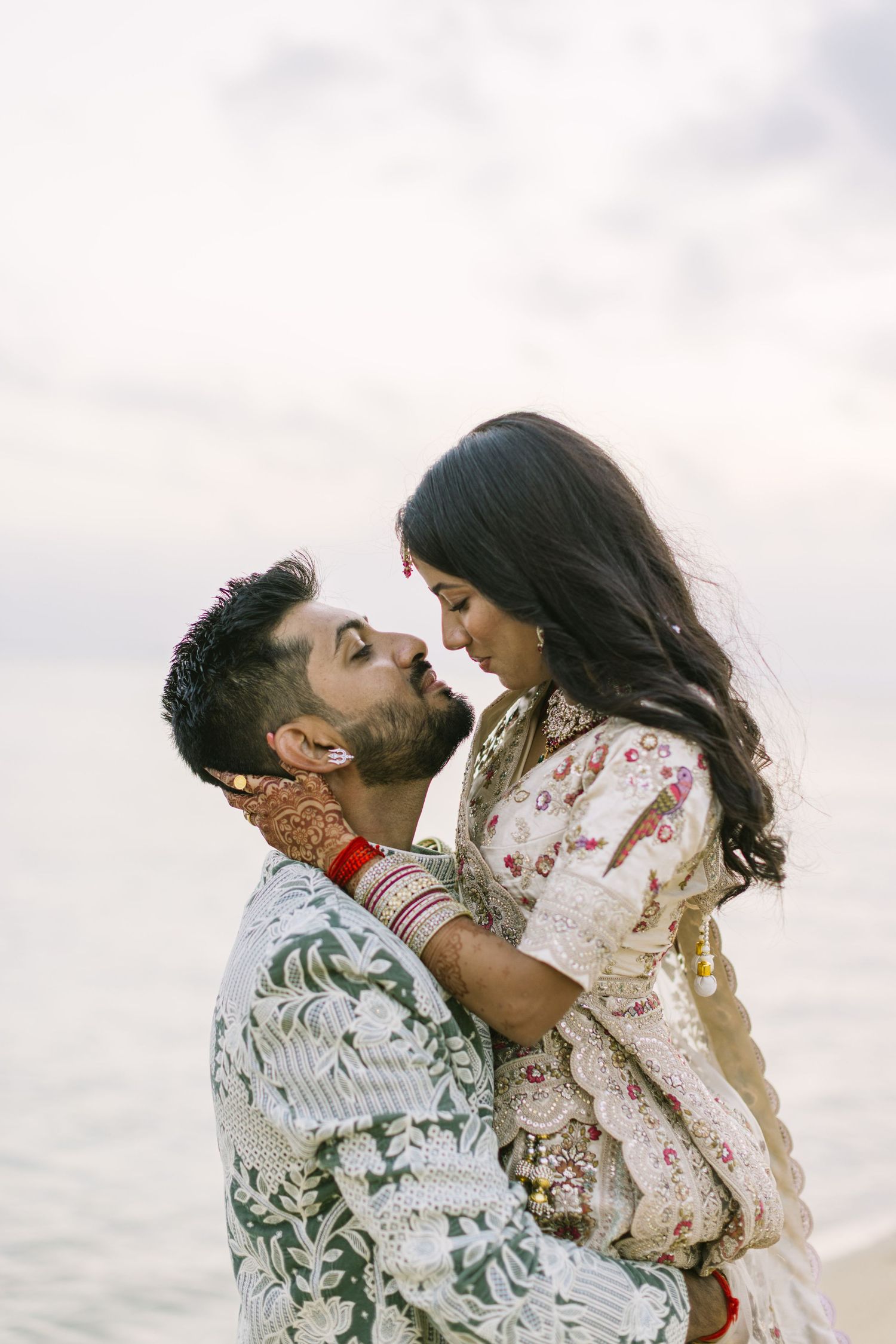 An Indian groom lifts his bride as they share an intimate moment near the ocean in traditional wedding attire.