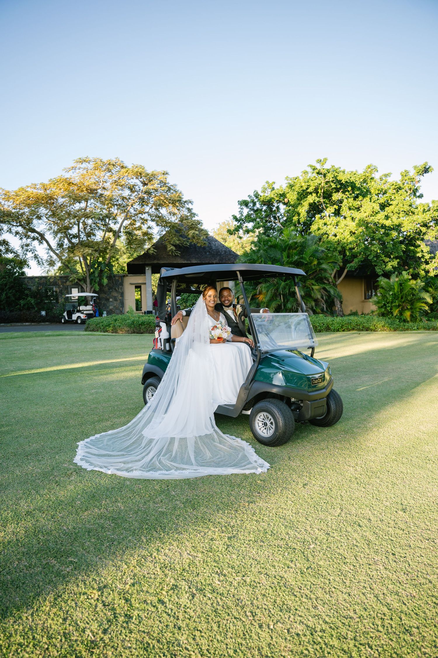 Bride and groom pose beside green golf cart on manicured lawn at sunset venue.