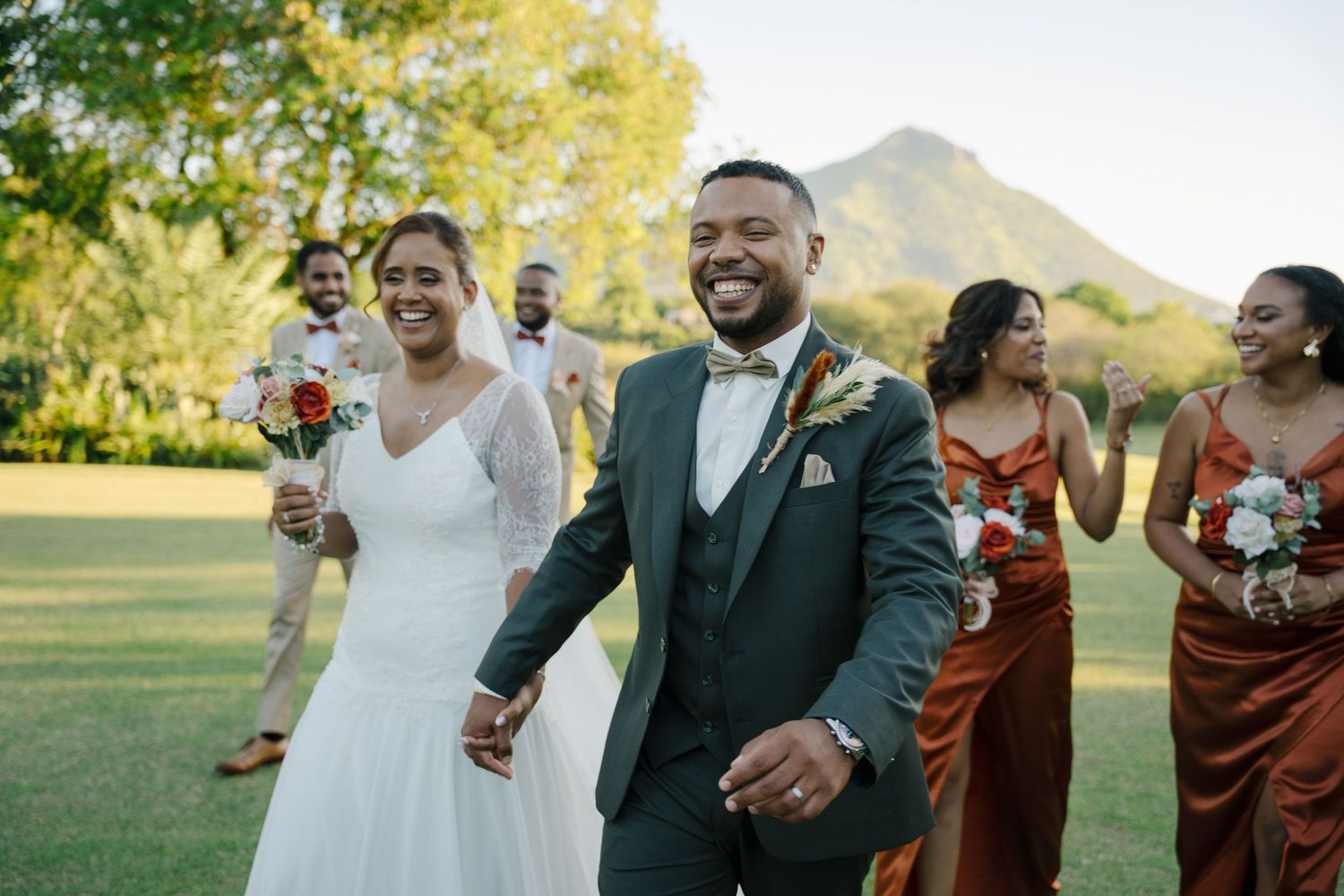 Smiling bride and groom lead joyful wedding party across sunlit lawn with mountain view.
