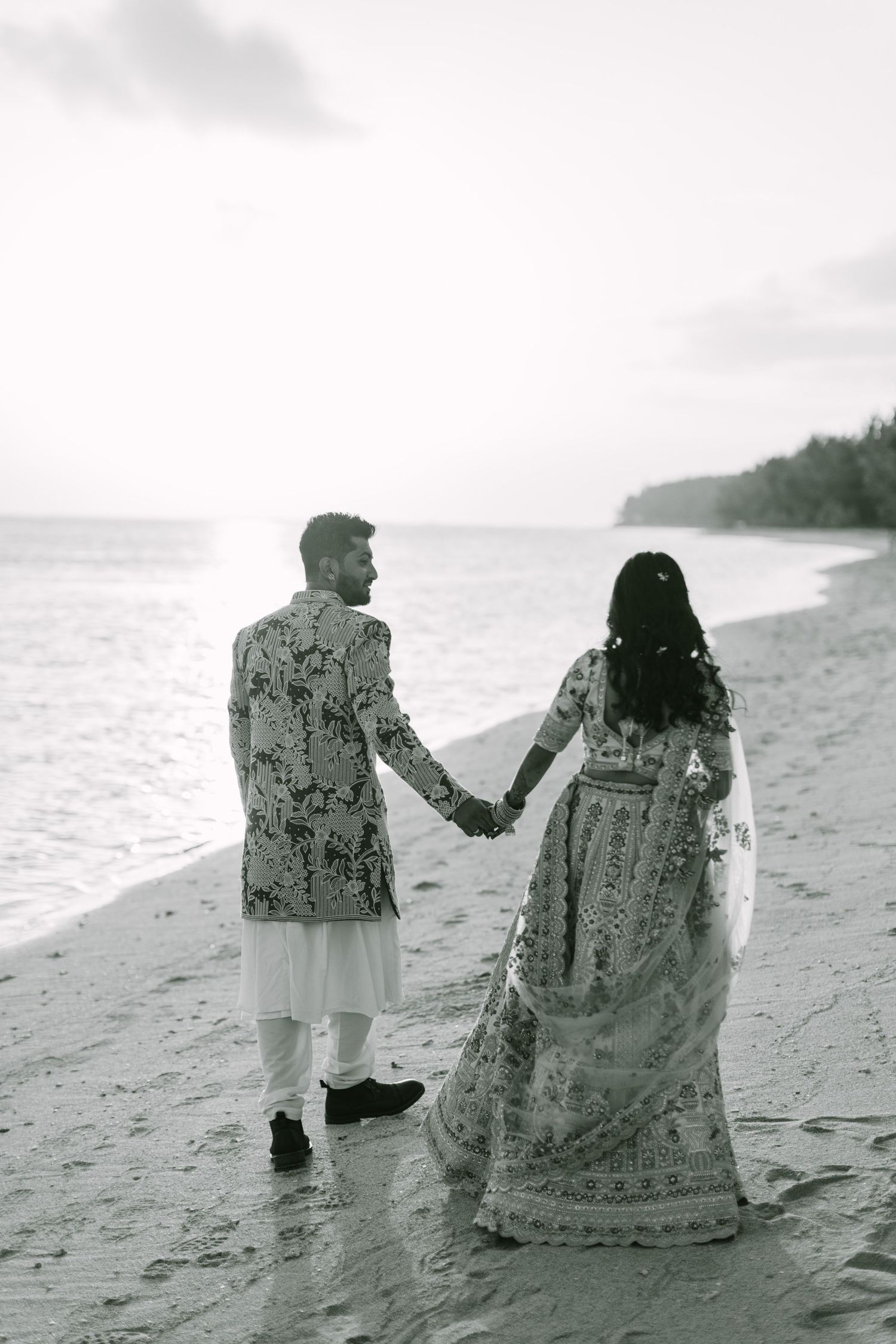 A black and white photo of an Indian wedding couple holding hands while walking along a peaceful beach shoreline.