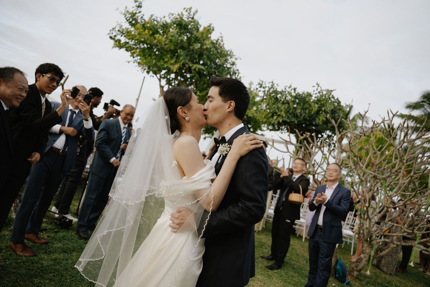 A bride and groom share their first kiss at an outdoor wedding ceremony surrounded by guests and lush greenery.