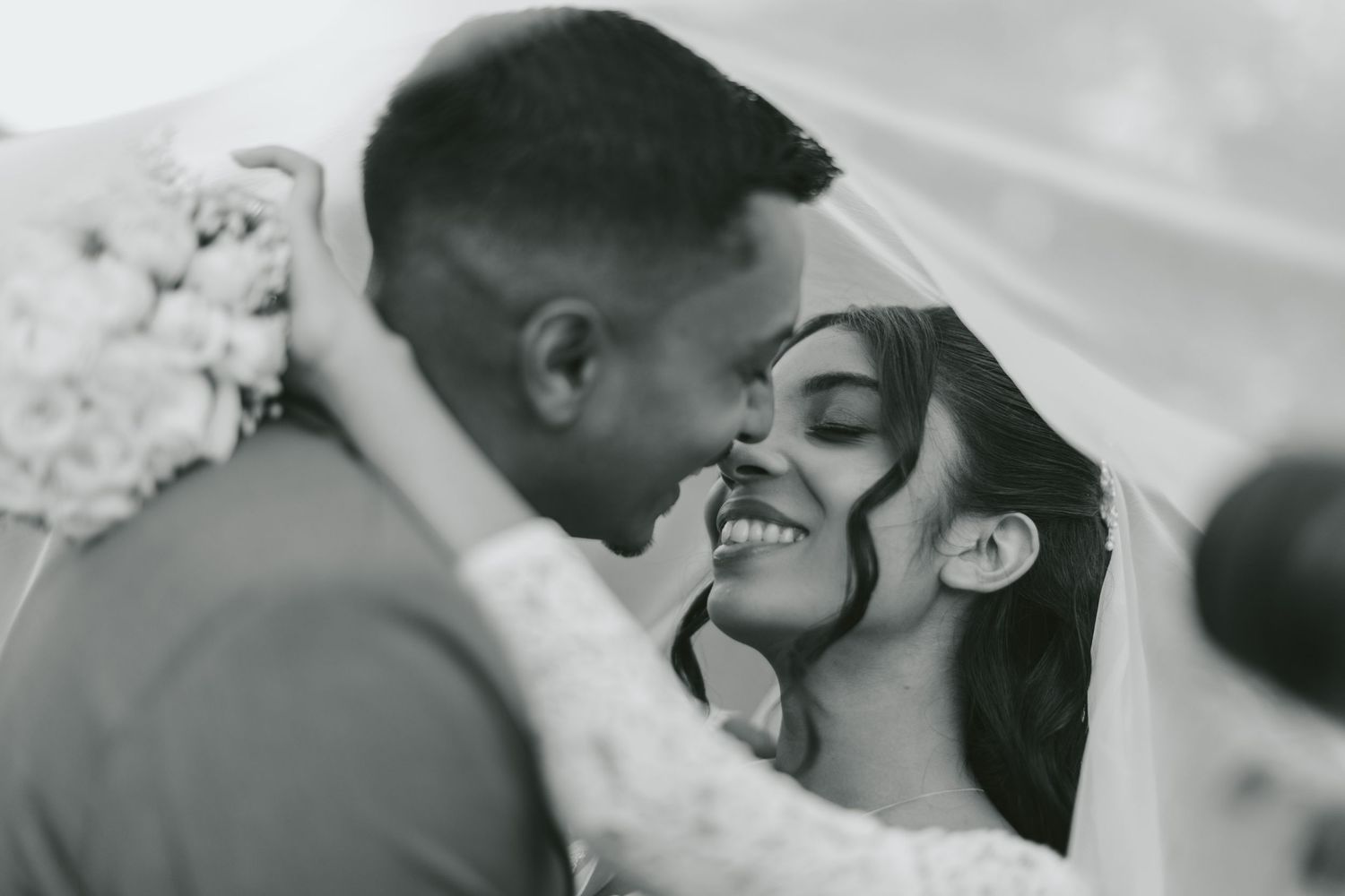 Black and white photo of smiling bride under veil, groom leaning in for a kiss, holding floral bouquet.