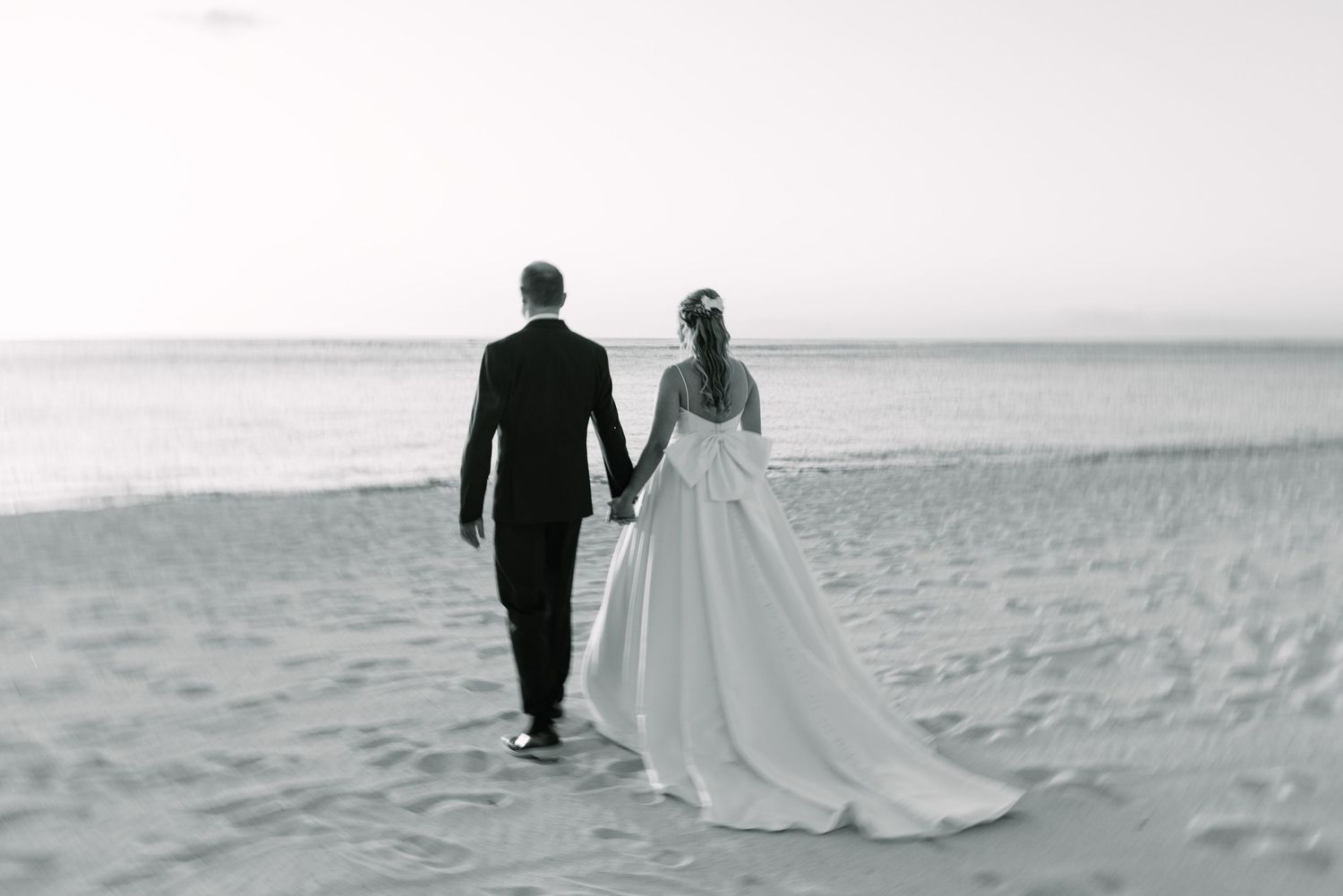 Bride and groom walking hand in hand along a sandy beach in black and white wedding photo.