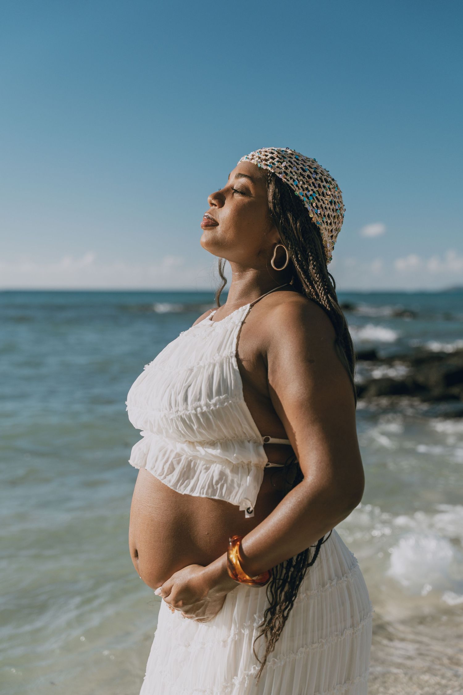 Pregnant woman in white crop top and skirt poses on a sunny beach with ocean waves in the background.