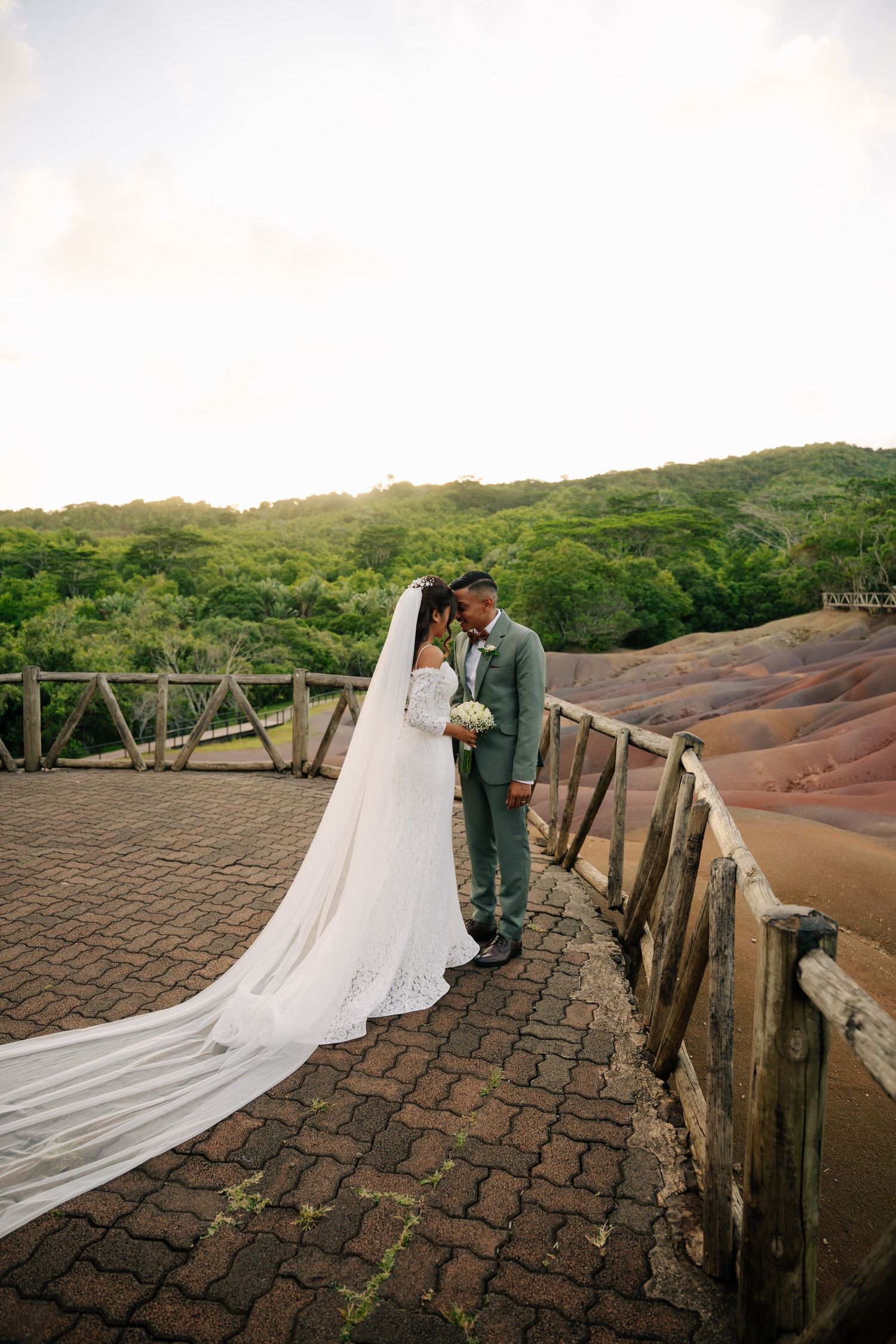 Bride and groom sharing a kiss on a scenic brick pathway with lush green hills and dramatic sky behind them.