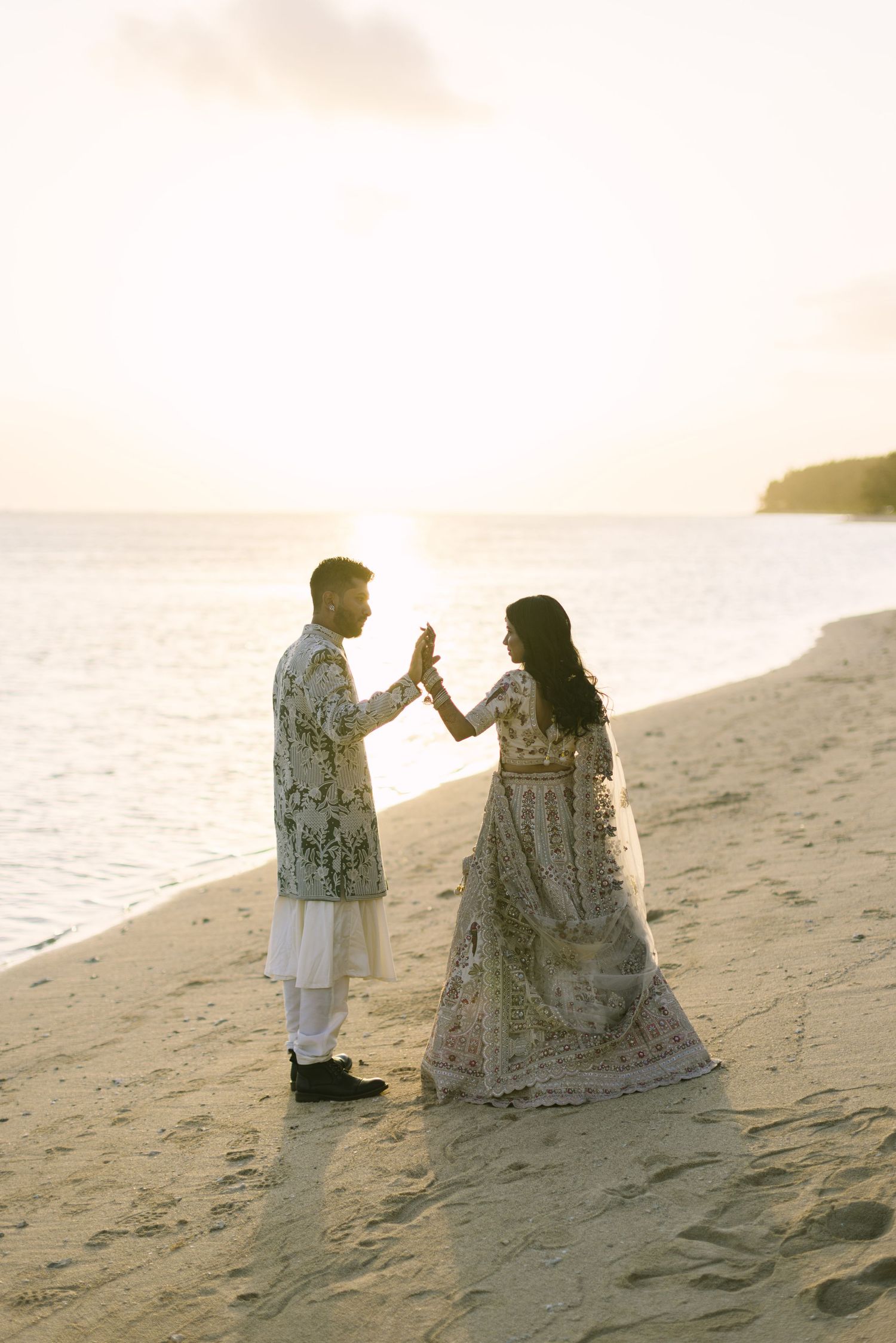 An Indian bride and groom touch hands at sunset on a sandy beach in ornate traditional wedding clothing.