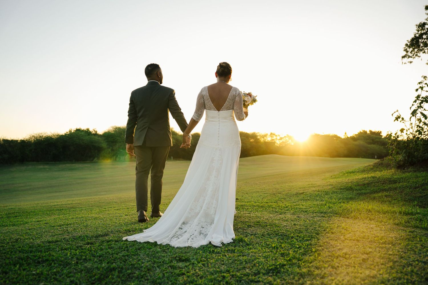 Bride and groom walk hand in hand toward glowing sunset on lush green lawn.
