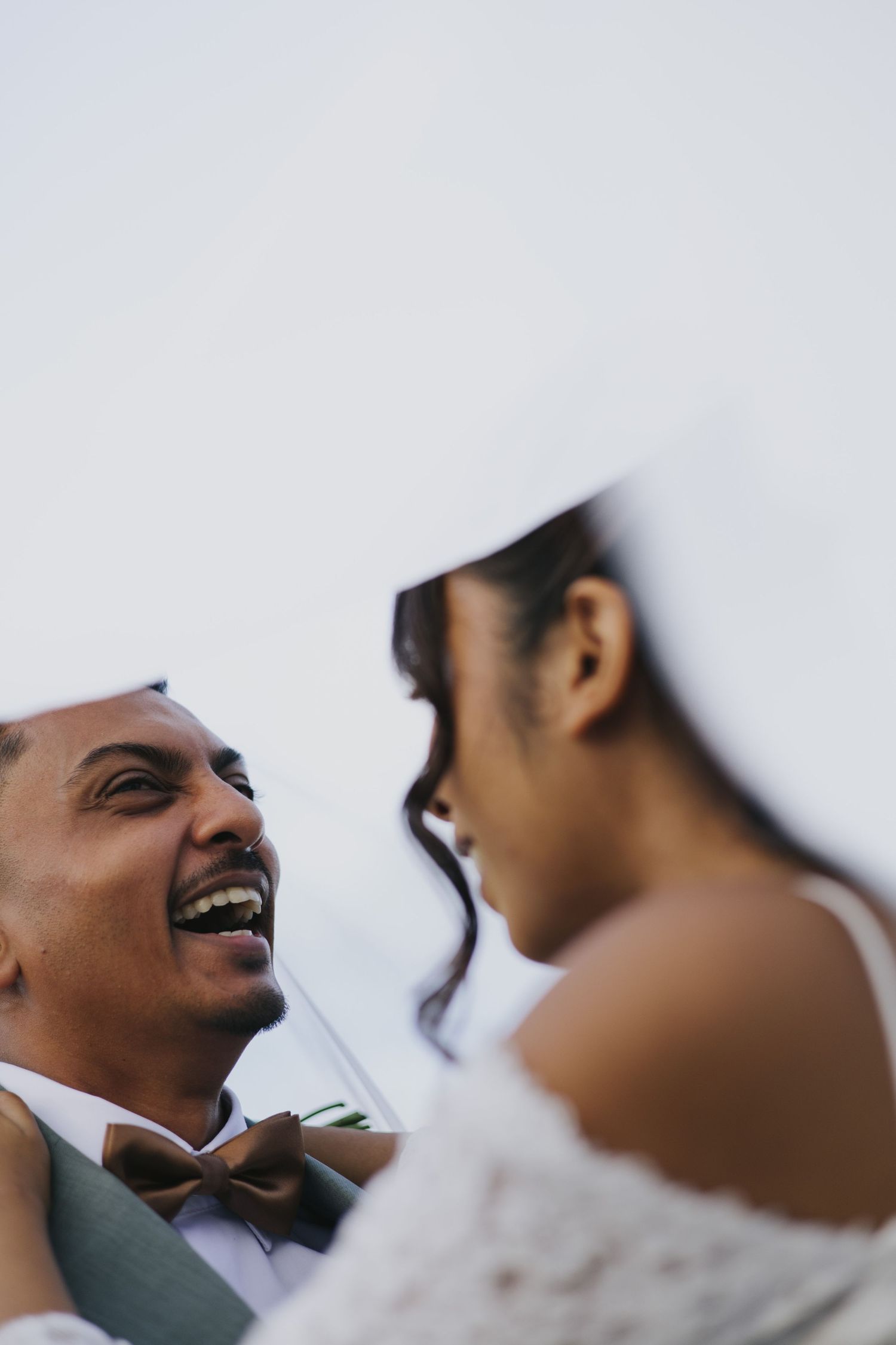 Joyful groom laughing with bride in white lace dress and veil during outdoor wedding ceremony.