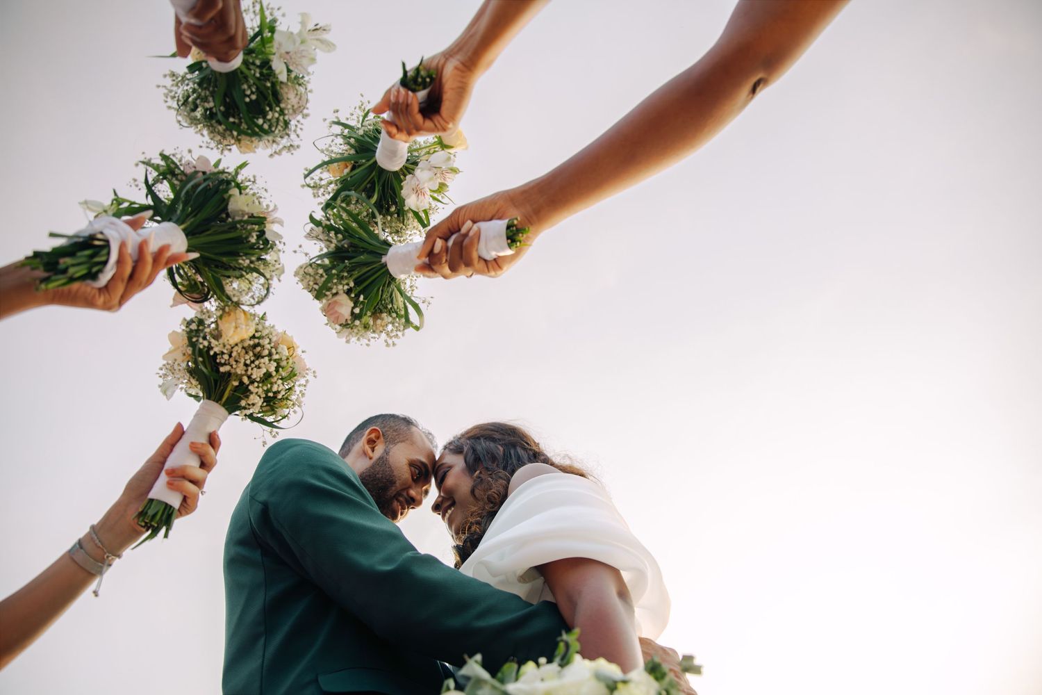 Wedding party raising bouquets over bride and groom touching foreheads in joyful celebration.