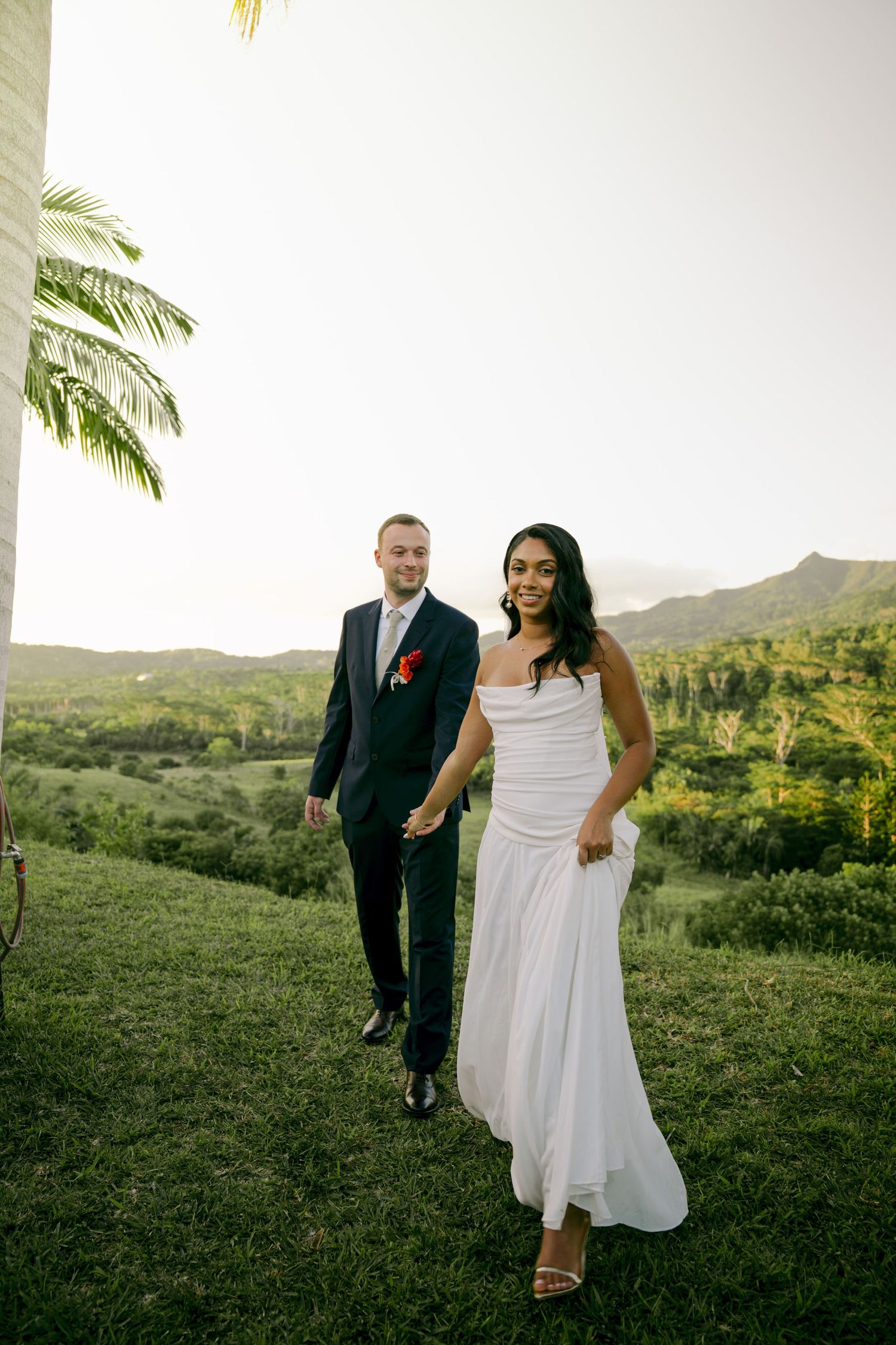 Bride in white gown and groom in dark suit walking hand in hand on green hillside at sunset.