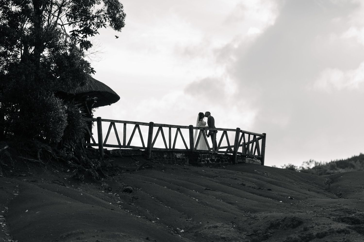 Black and white silhouette of bride and groom kissing on a wooden overlook platform against a cloudy sky.