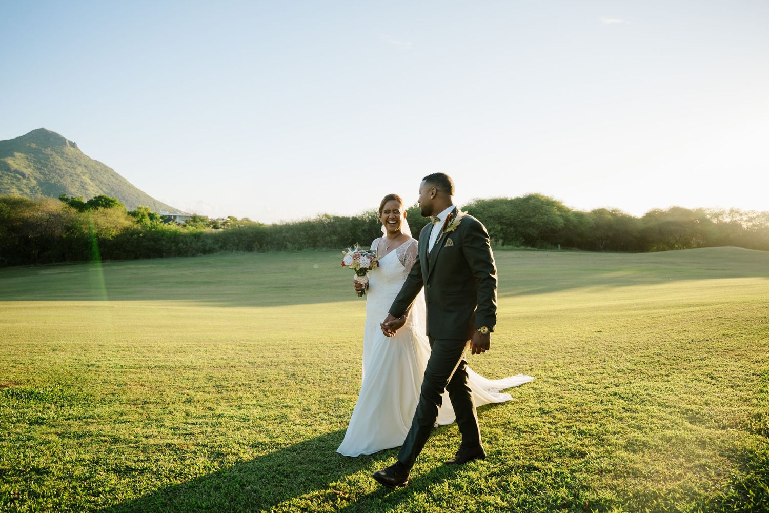 Bride and groom walk hand in hand across green field with mountain in background.