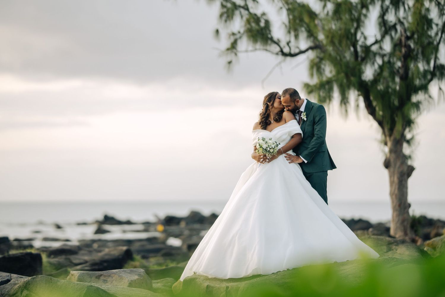 Bride and groom share romantic kiss by rocky ocean shore surrounded by tropical trees.