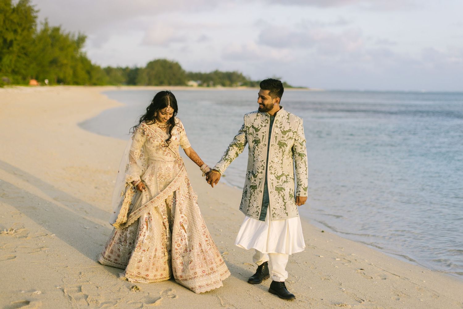 An Indian bride and groom in ornate traditional attire walk hand in hand along a tropical sandy beach.
