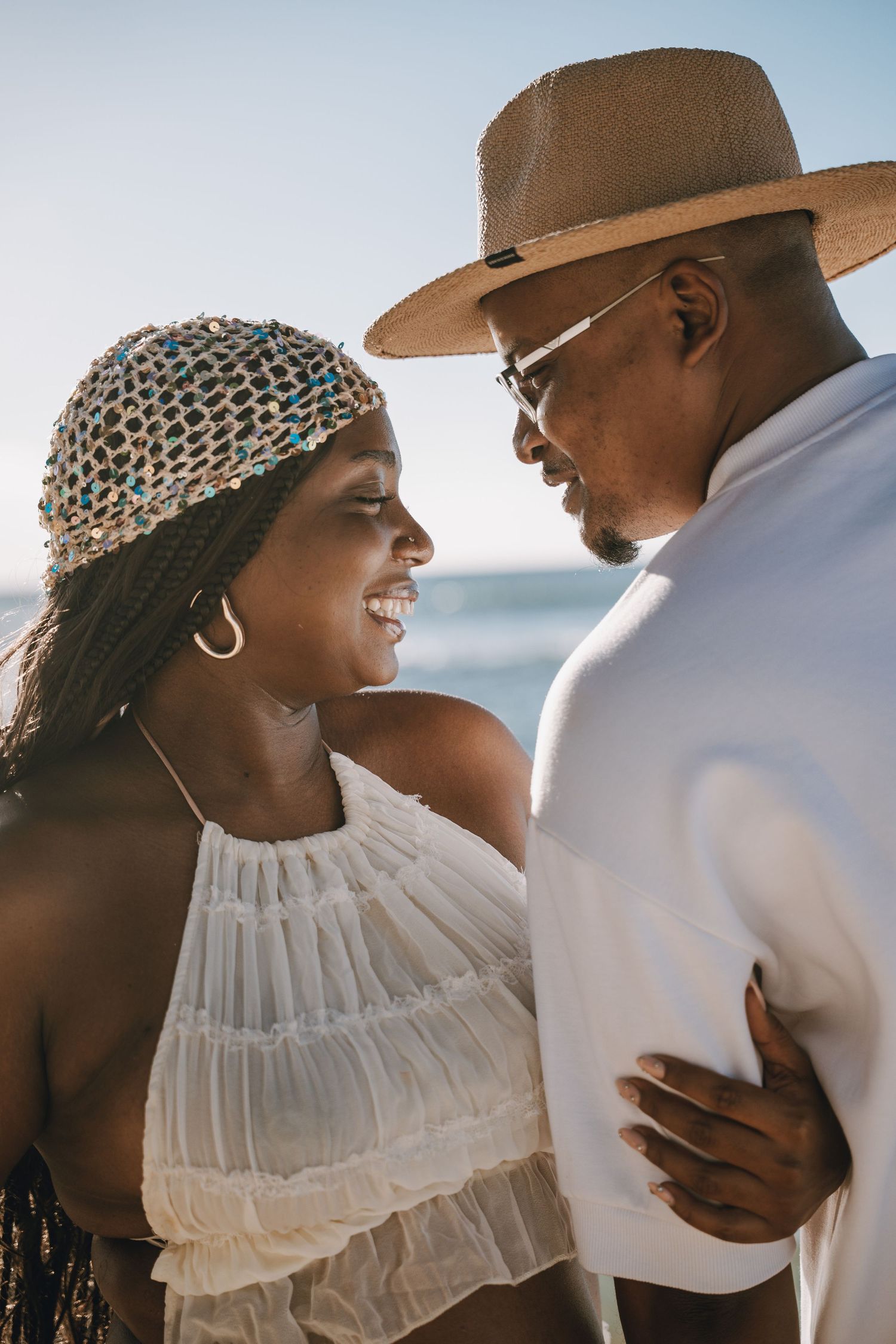Smiling couple sharing a romantic moment on a sunny beach, woman in white halter top and beaded headpiece, man in straw hat.