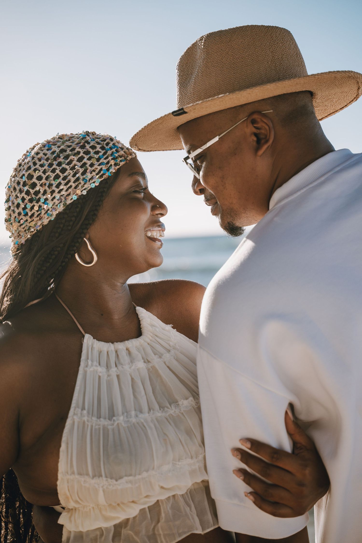 Happy couple embracing on sunny beach, woman in white top with braids, man in straw hat and white shirt.