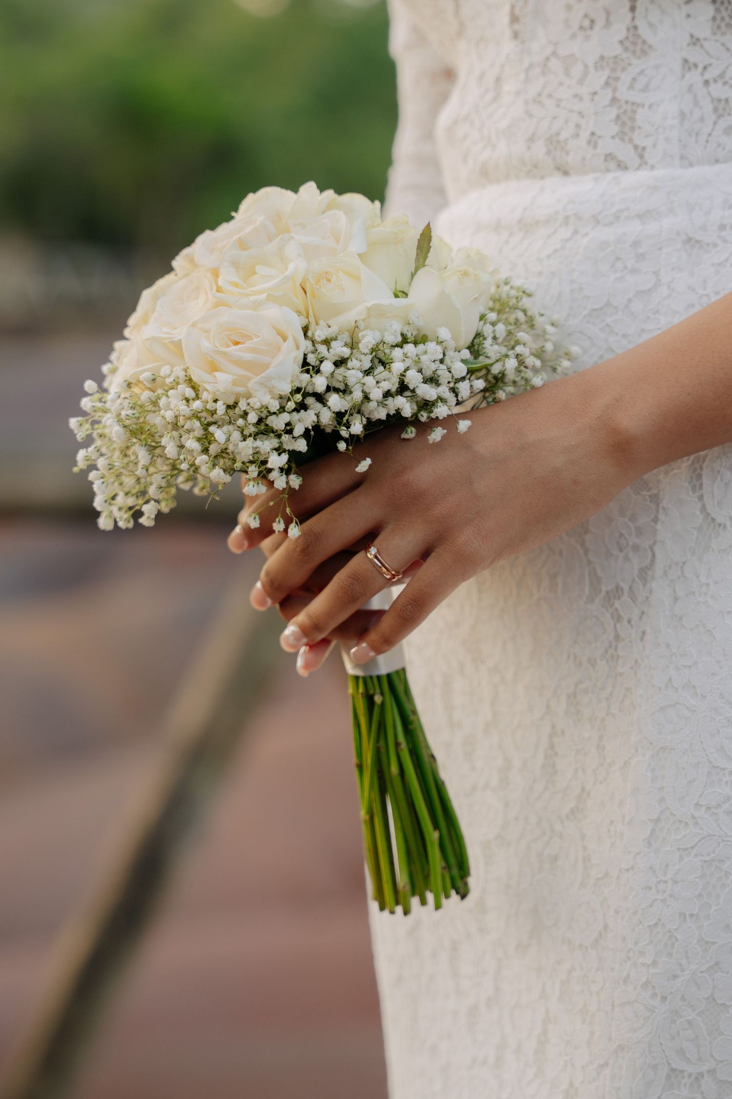 Bride holding elegant white rose and baby's breath bouquet, wearing gold wedding ring, in lace dress.