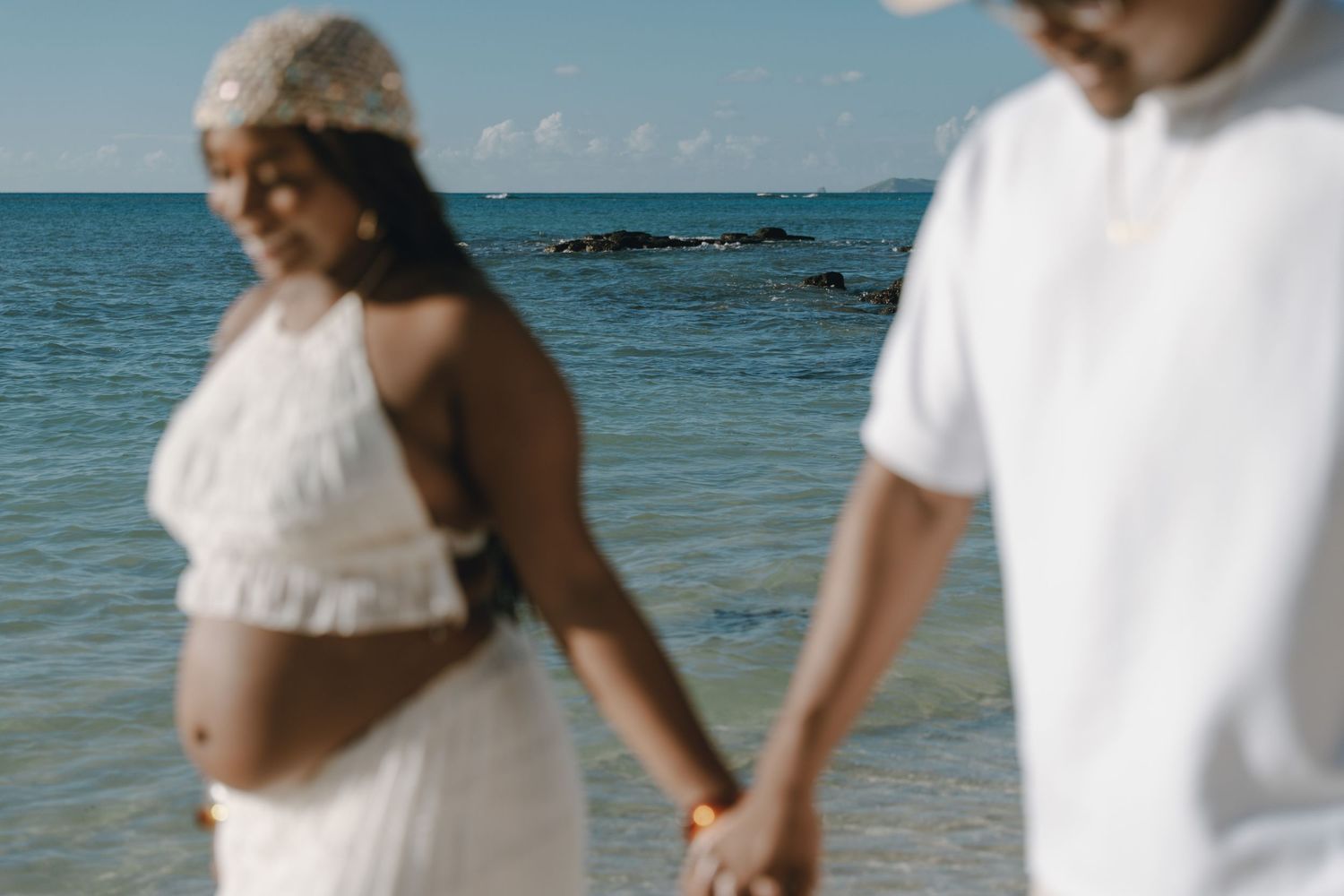 Pregnant woman in white dress holds hands with partner on a tropical beach with turquoise ocean in background.