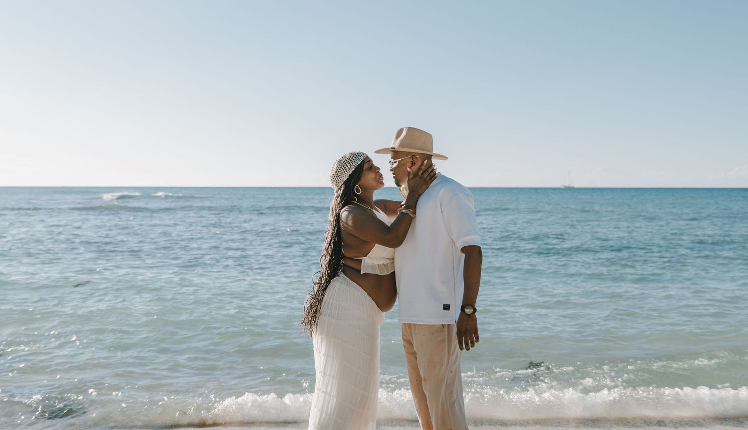 Couple embracing romantically on a beach shoreline, woman in white maternity dress, man in white outfit and wide-brim hat.