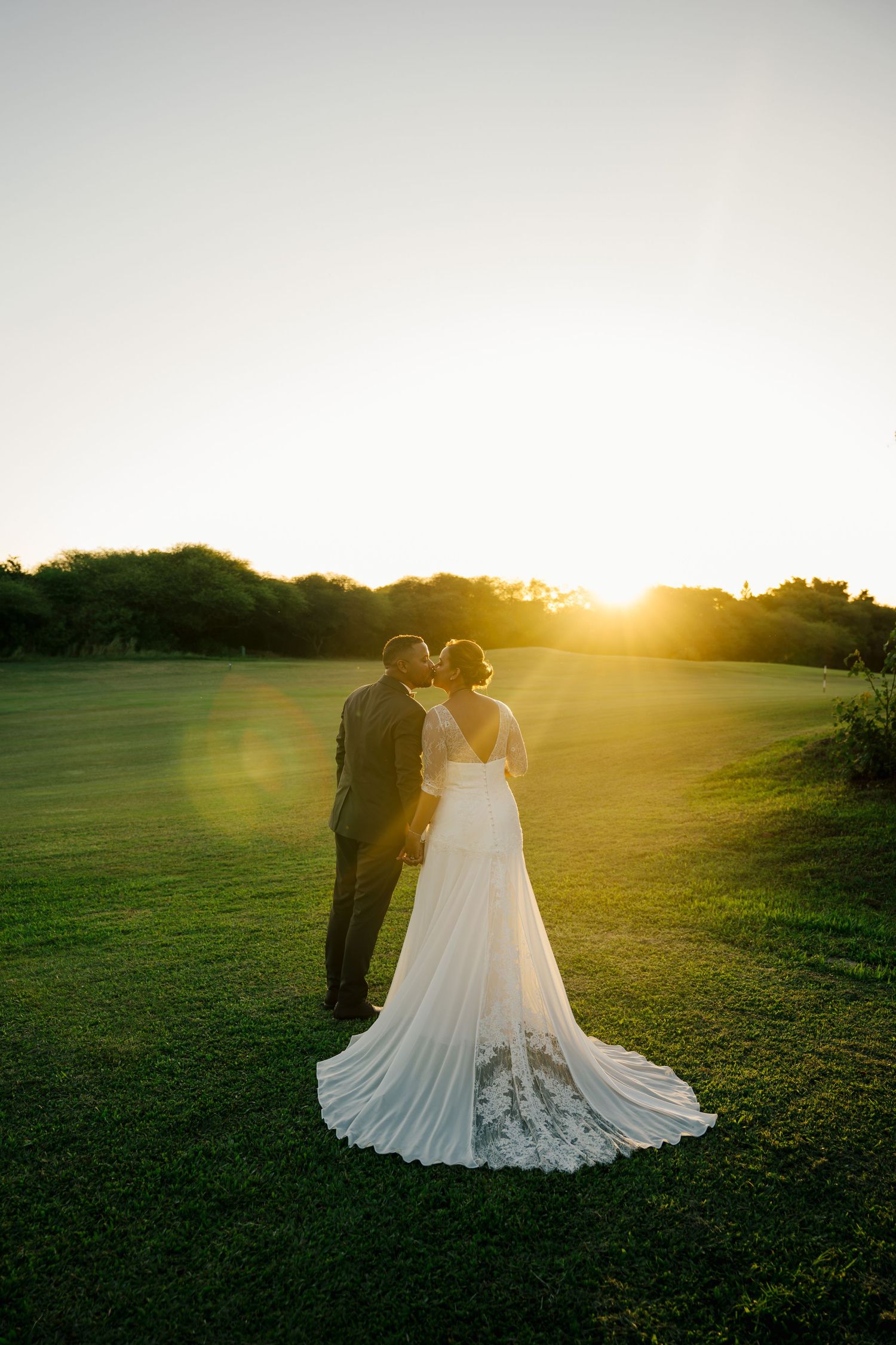 Couple shares sunset kiss on green lawn, bride's lace train flowing behind her.