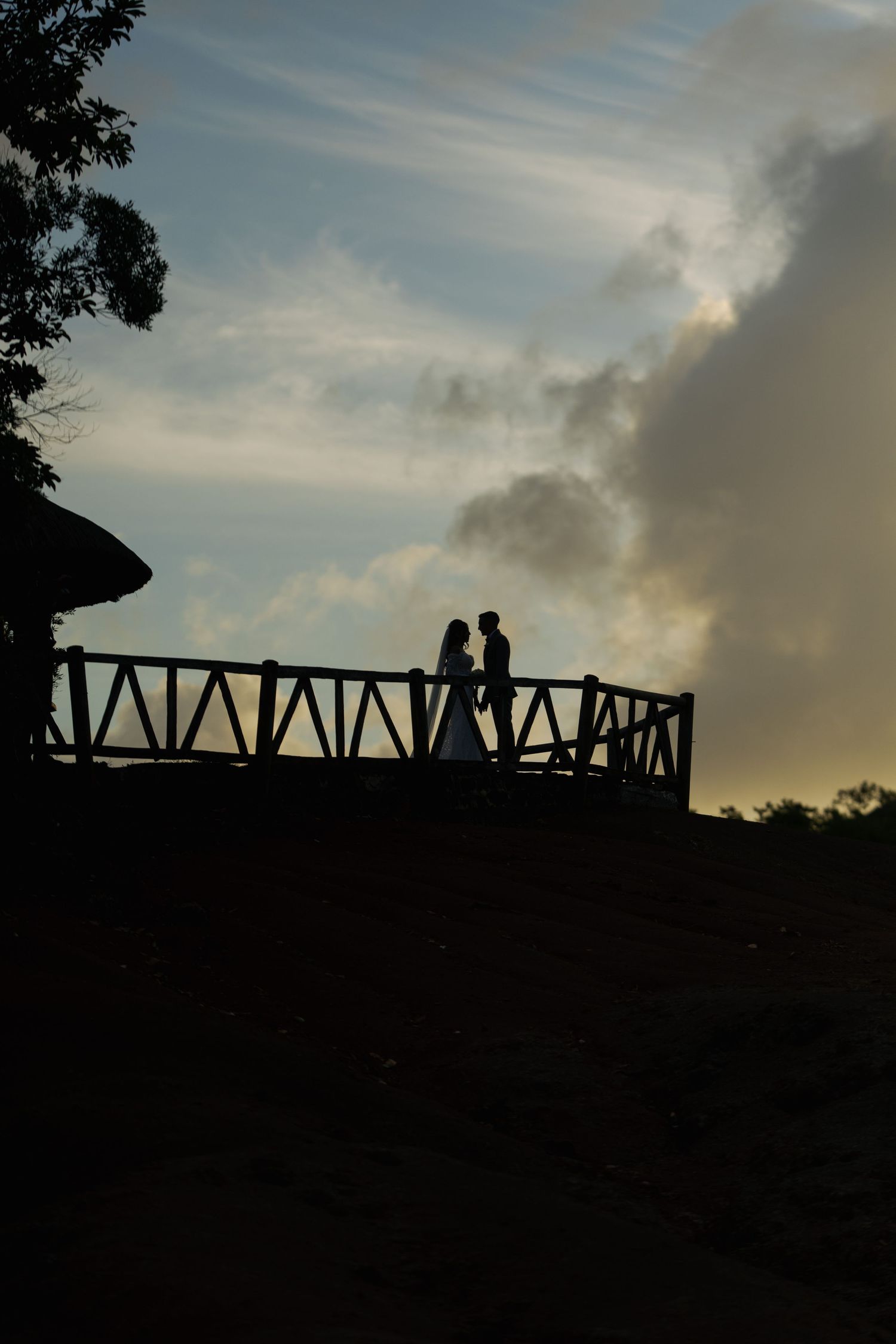 Romantic silhouette of bride and groom facing each other on a wooden deck overlook at dramatic sunset.