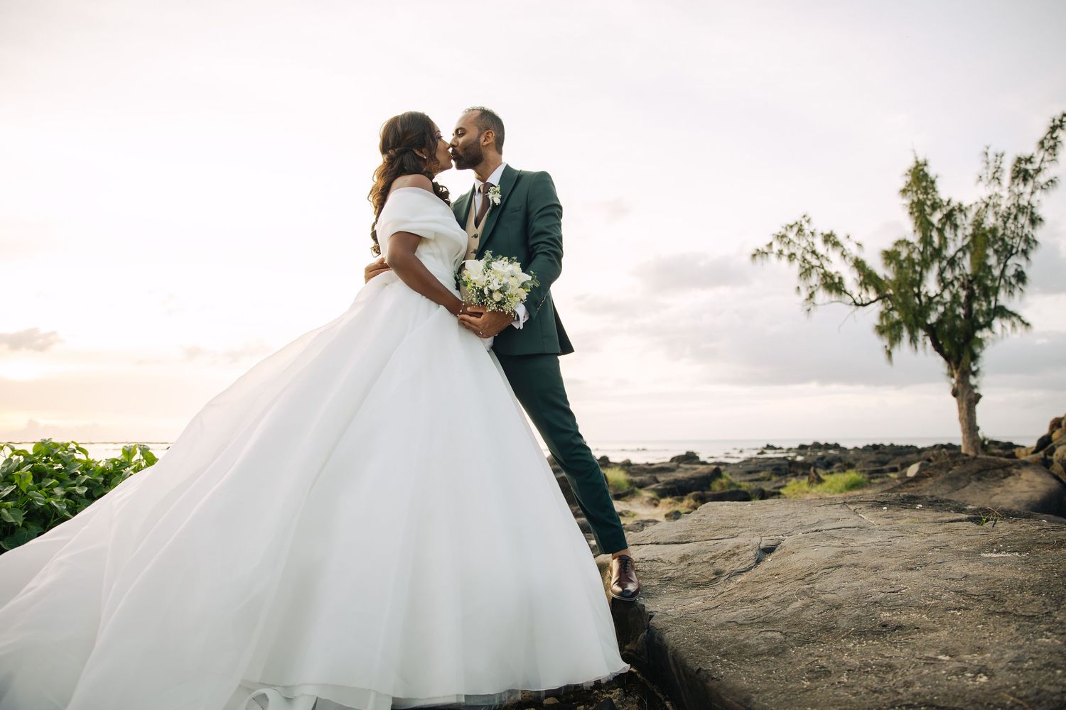 A bride in a voluminous white ball gown and groom in a teal suit kiss on a rocky scenic overlook.