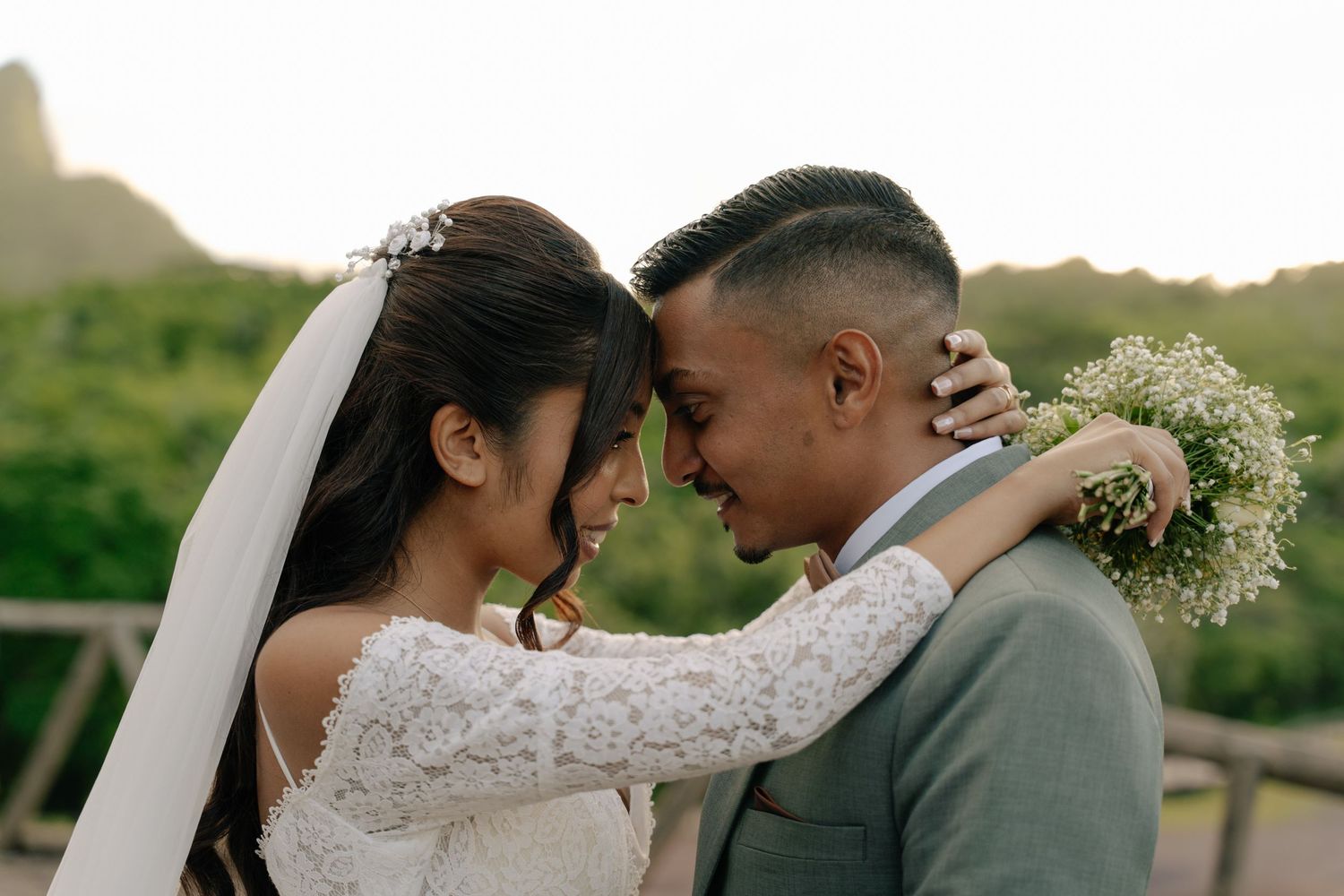 Bride and groom touching foreheads tenderly, smiling, with lush green hills in the background.