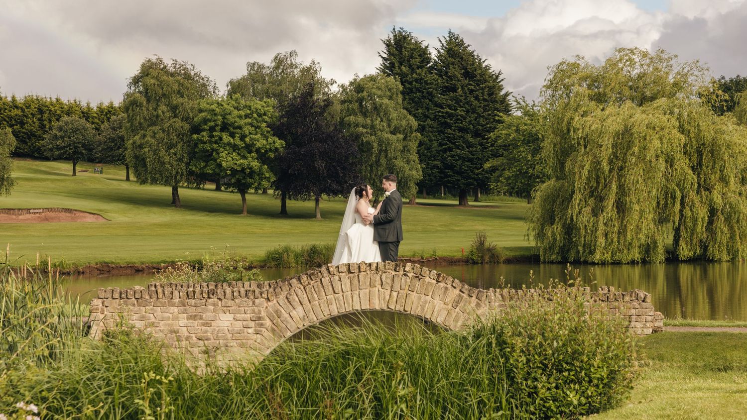 Cortney and Dan standing together on a stone bridge over the lake at Windmill Village Hotel in Coventry.