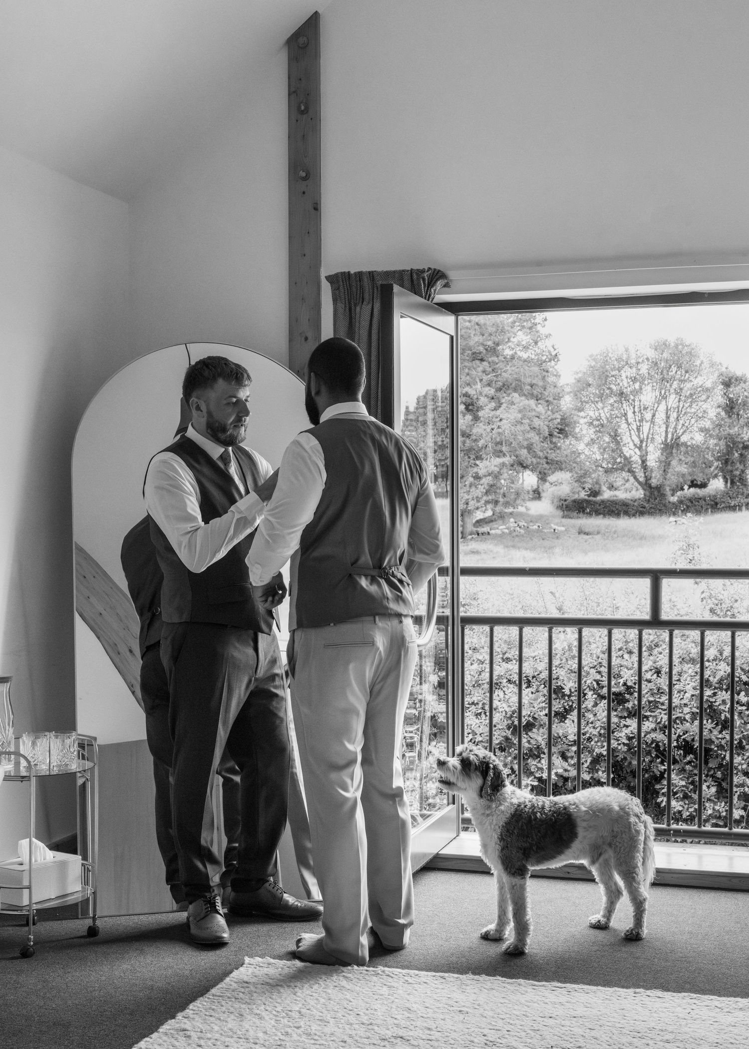 Jack helps Aaron adjust his wedding waistcoat while their dog Milo watches beside the open balcony doors at Rock Village Hall.