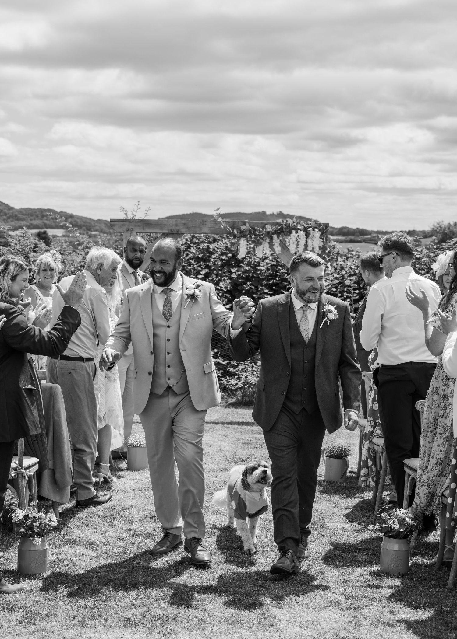 Aaron and Jack walk hand in hand down the outdoor aisle with Milo as guests applaud at Rock Village Hall.