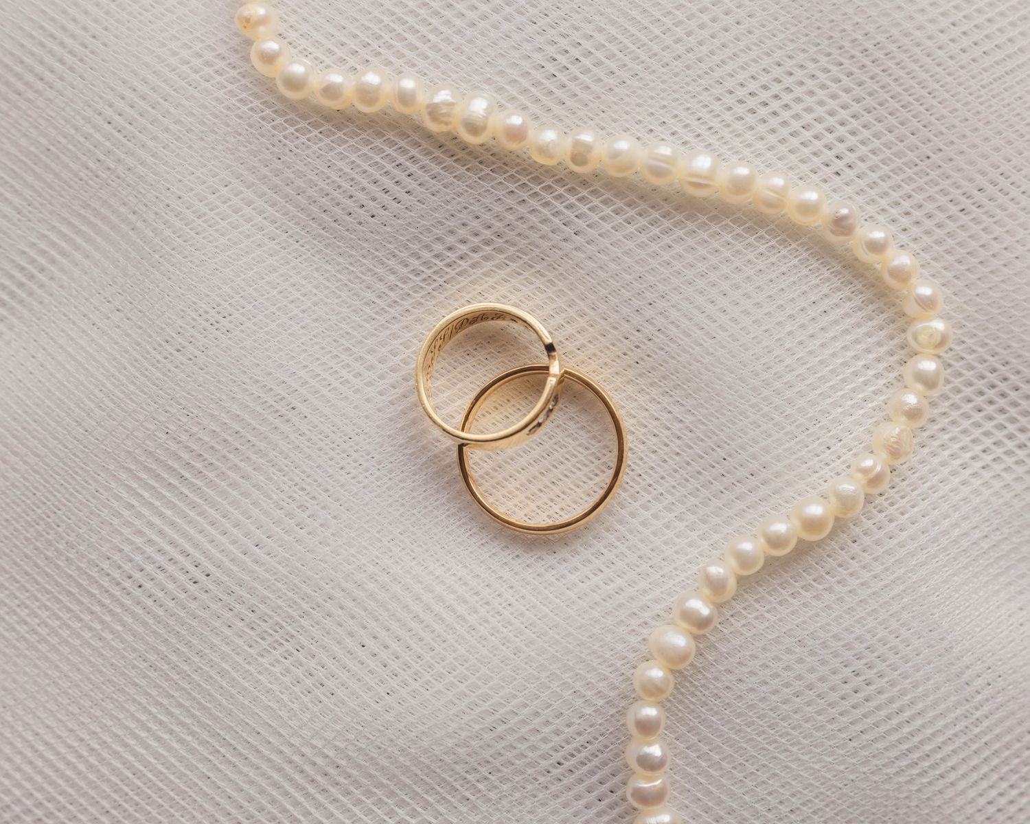 Two gold wedding rings and a pearl necklace on top of a white background, captured in a natural, romantic style by EvieSJPhotography.