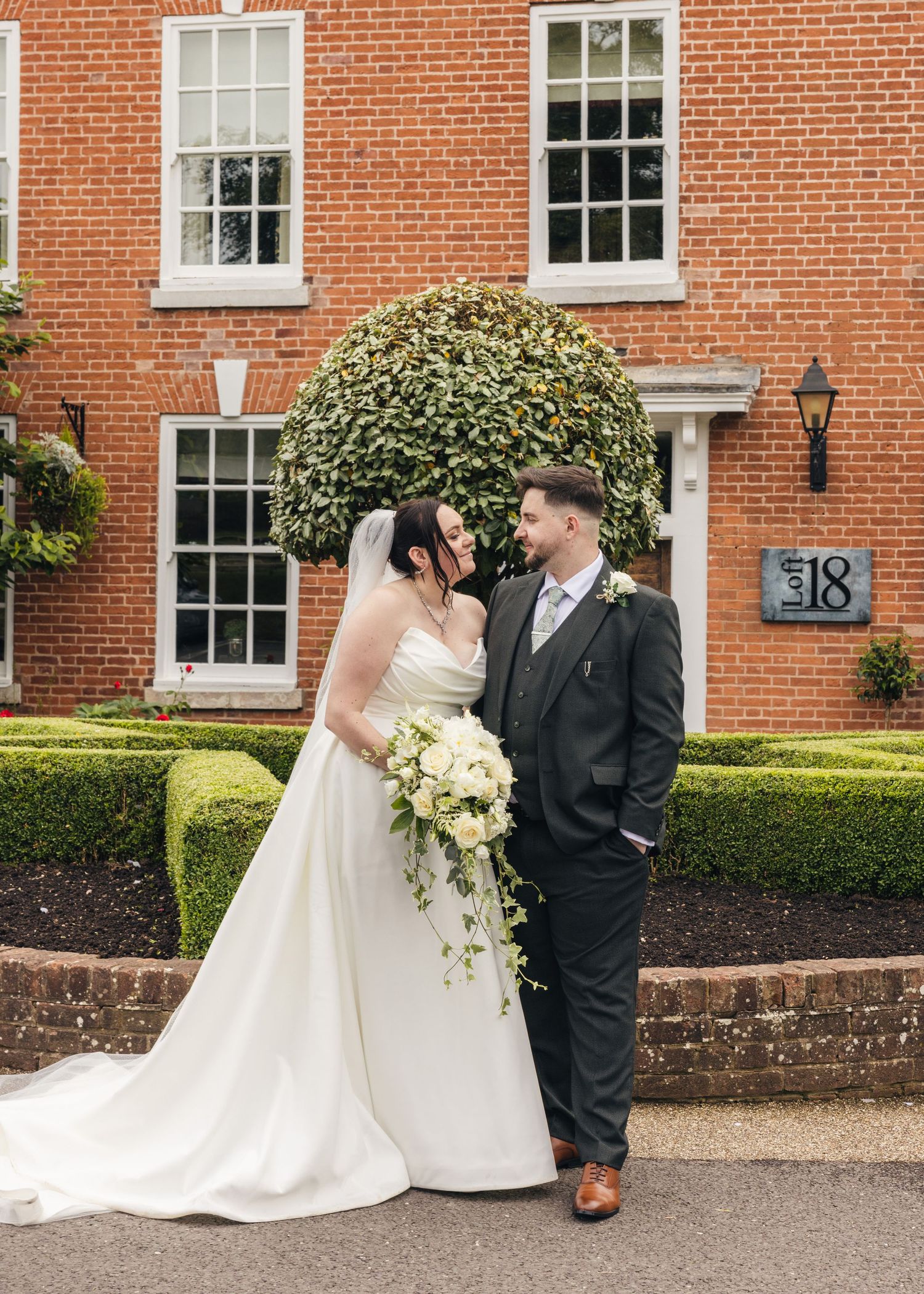 Cortney and Dan posing together in front of the red-brick buildings and gardens at their wedding venue.