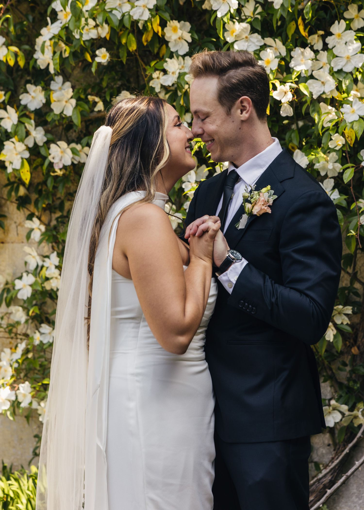 A bride and groom sharing a nose kiss in front of some flowers.
