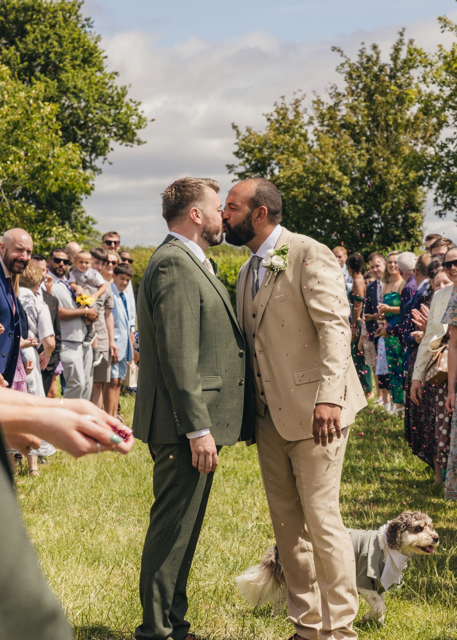 Aaron and Jack pause for a kiss with Milo beside them as guests throw colourful confetti outside Rock Village Hall.