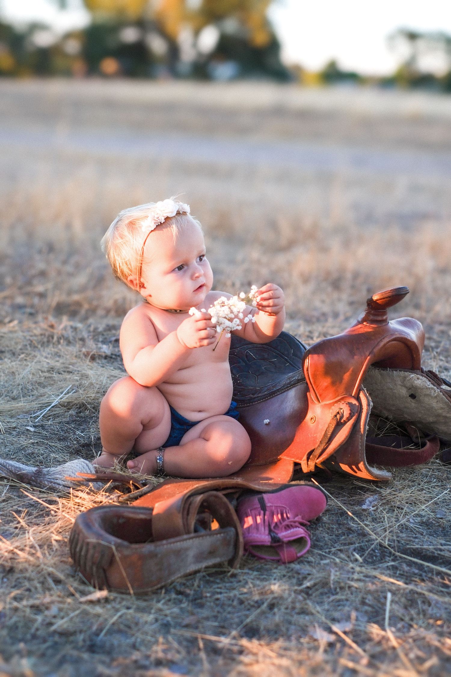 Baby in diaper and headband plays near leather saddle and boots on dry grass during sunset.