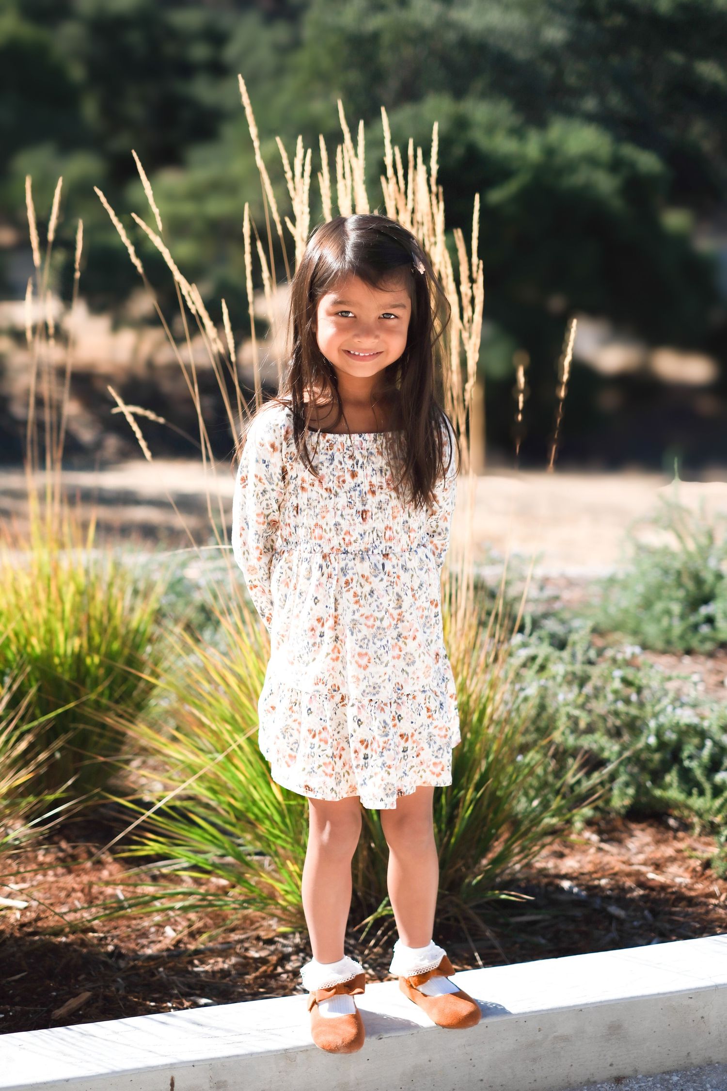 Young child in a floral dress standing in front of ornamental grasses in a garden setting.