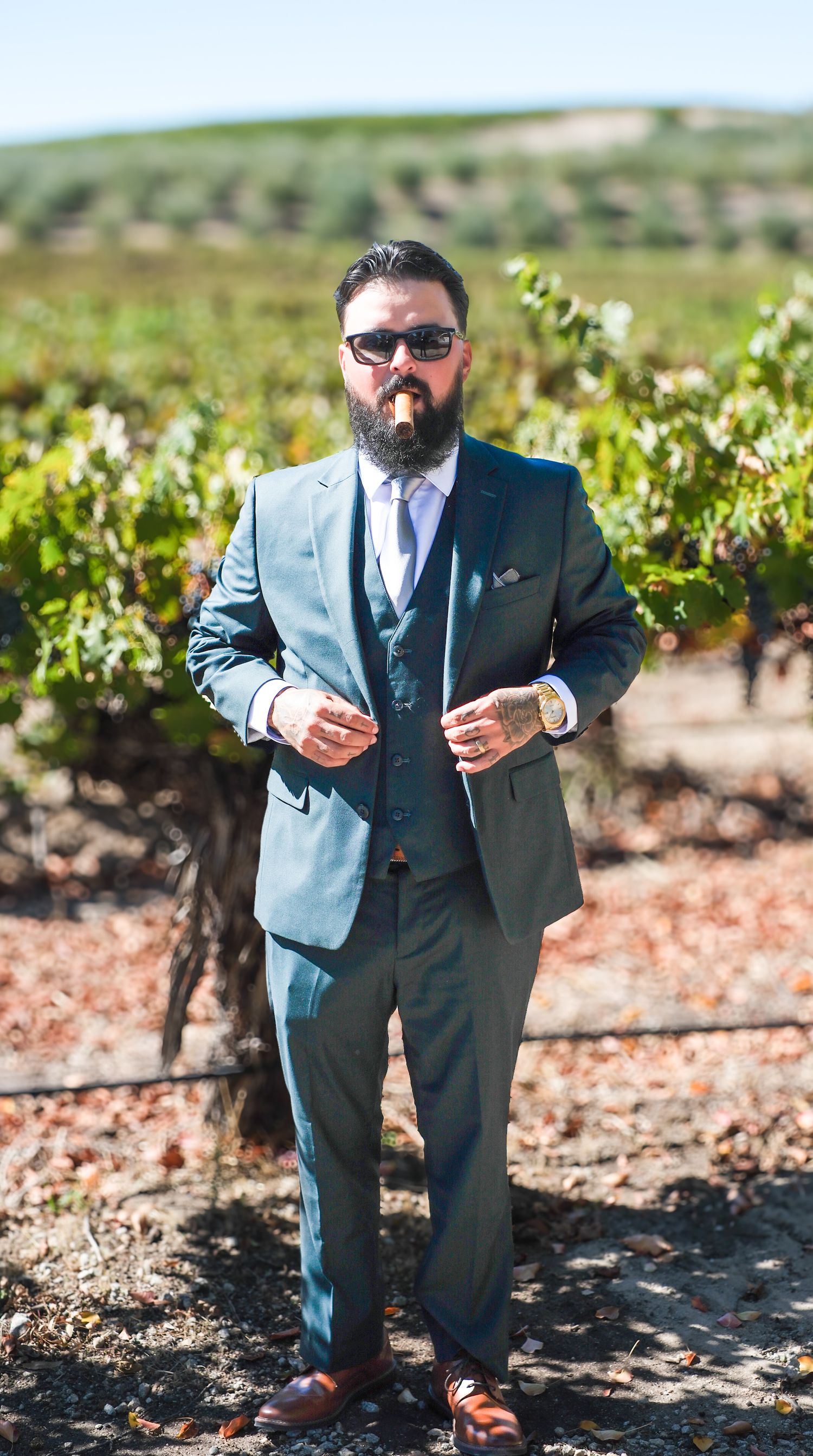 Groom in a light blue suit poses for creative portrait shots in a sunny vineyard setting.