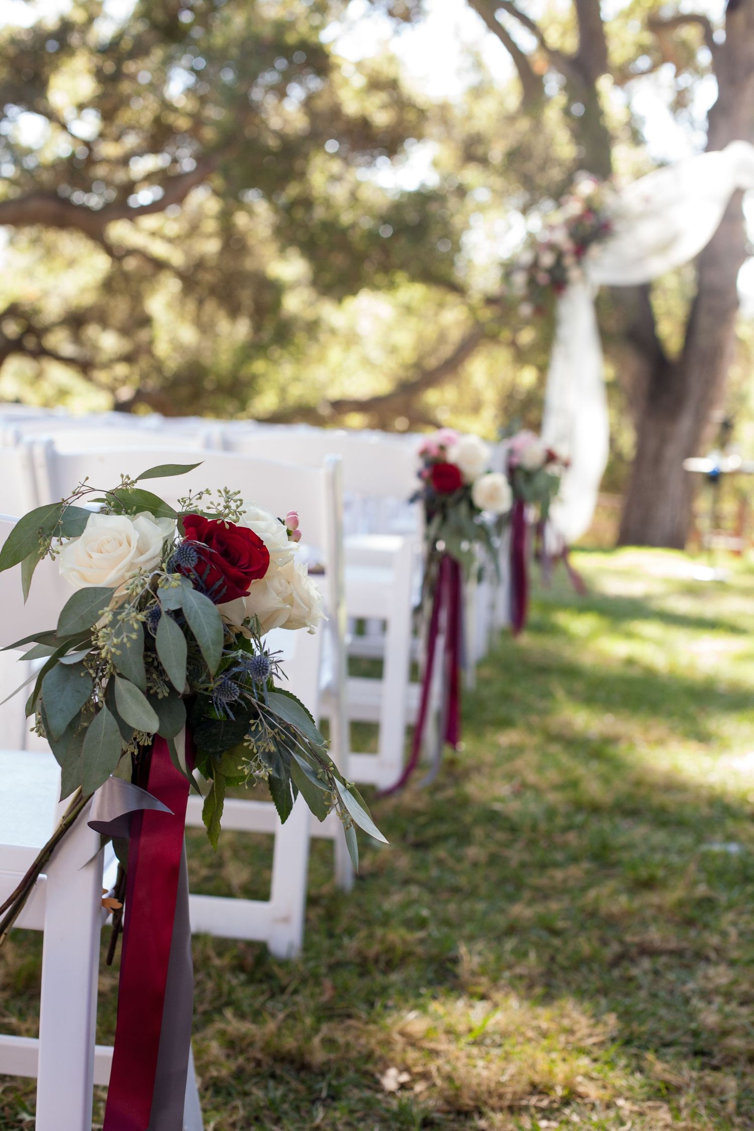White folding chairs decorated with burgundy ribbons and greenery line a wedding ceremony aisle outdoors.