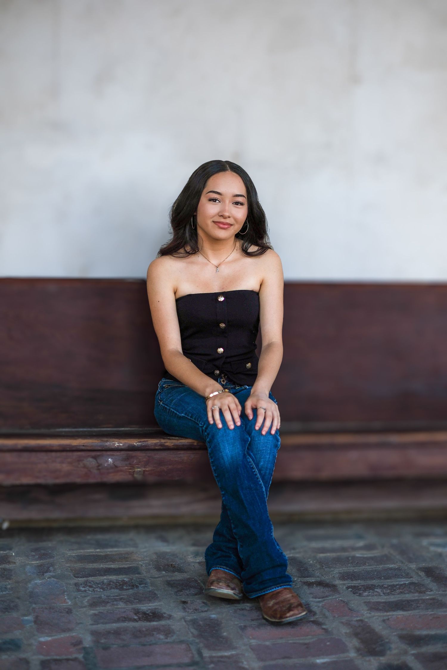 A person in a black strapless top and blue jeans sits casually on a bench against a textured wall.
