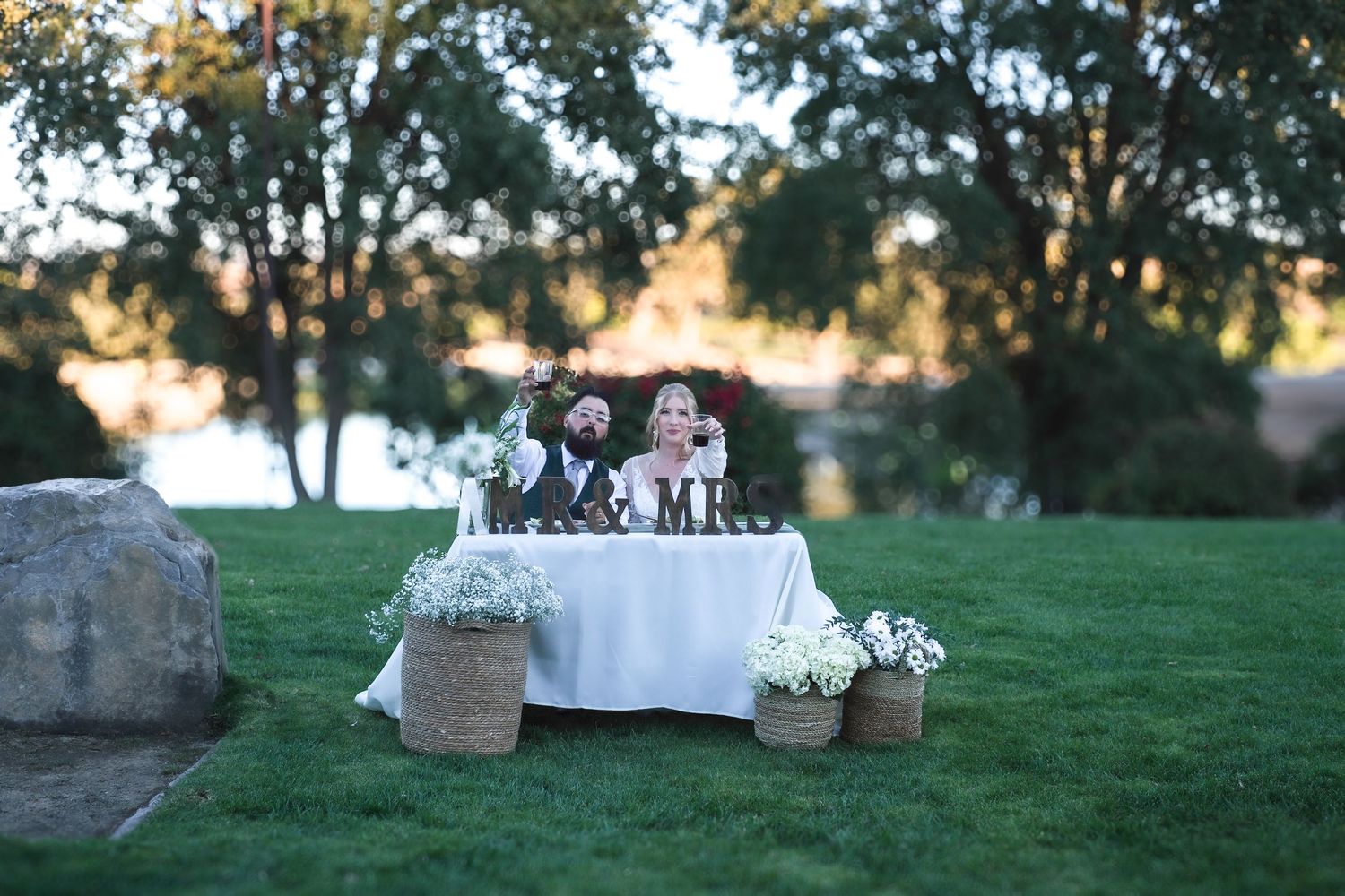Outdoor wedding reception setup with white linens and floral arrangements on a grassy hillside.