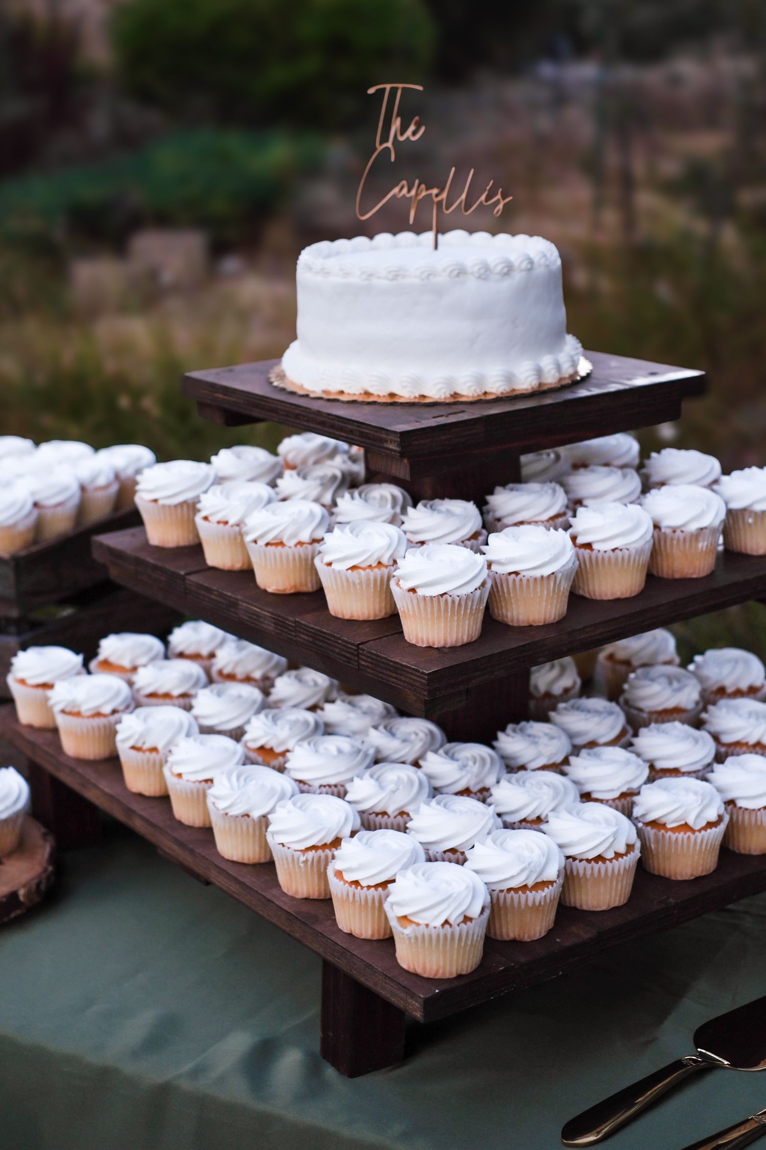 White frosted wedding cupcakes and cake displayed on tiered wooden stands at an outdoor reception.