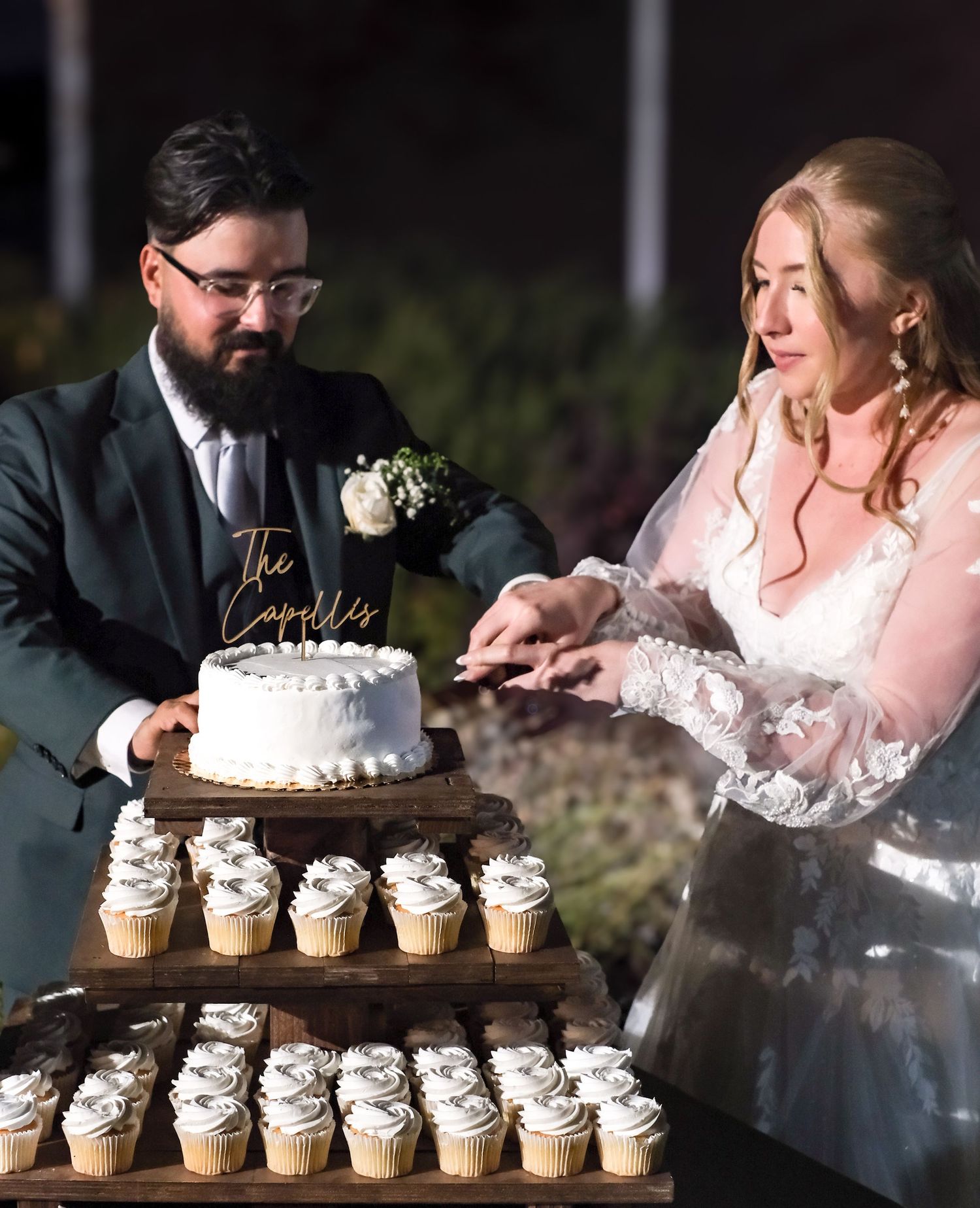 Wedding dessert display featuring a white tiered cake and cupcakes being served at an outdoor reception.