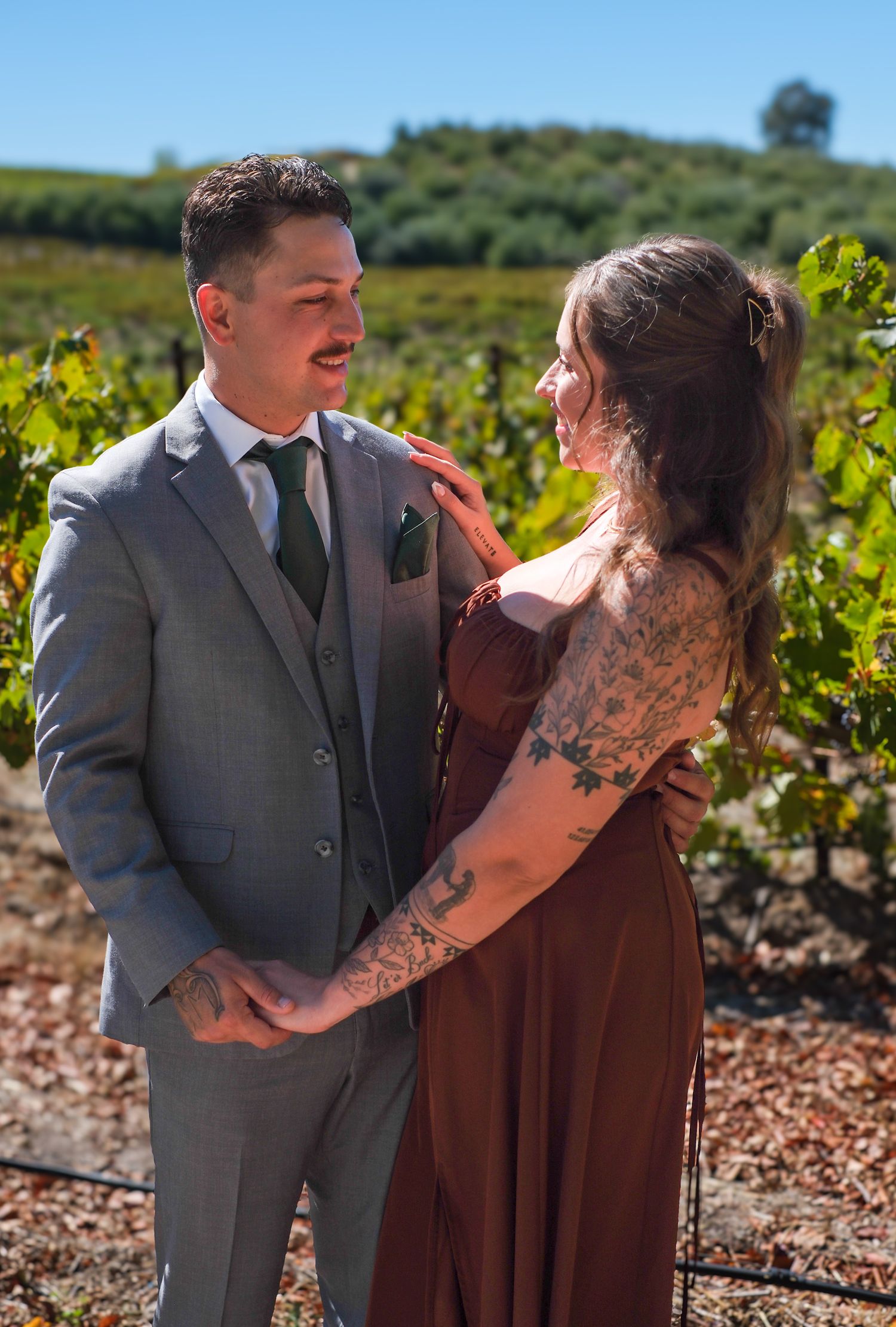 A couple embraces in a sunny vineyard, with rows of grapevines and hills in the background.
