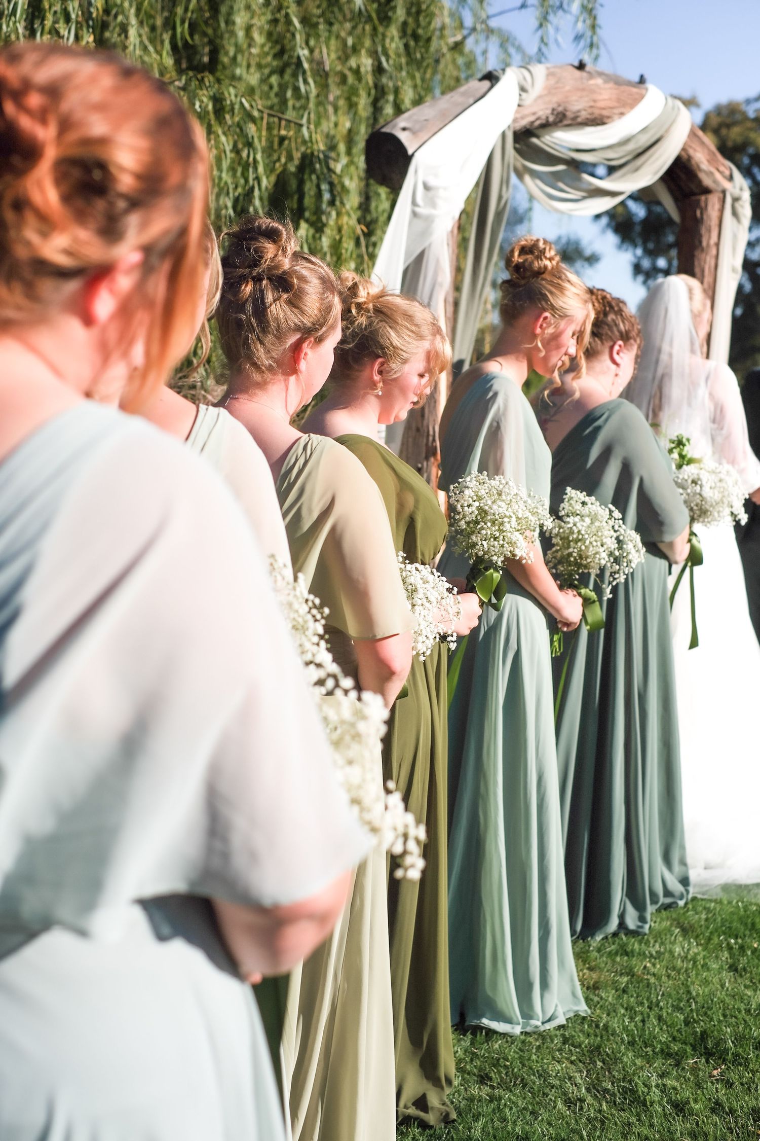 Bridesmaids in sage green dresses stand together during an outdoor wedding ceremony under a weeping willow tree.