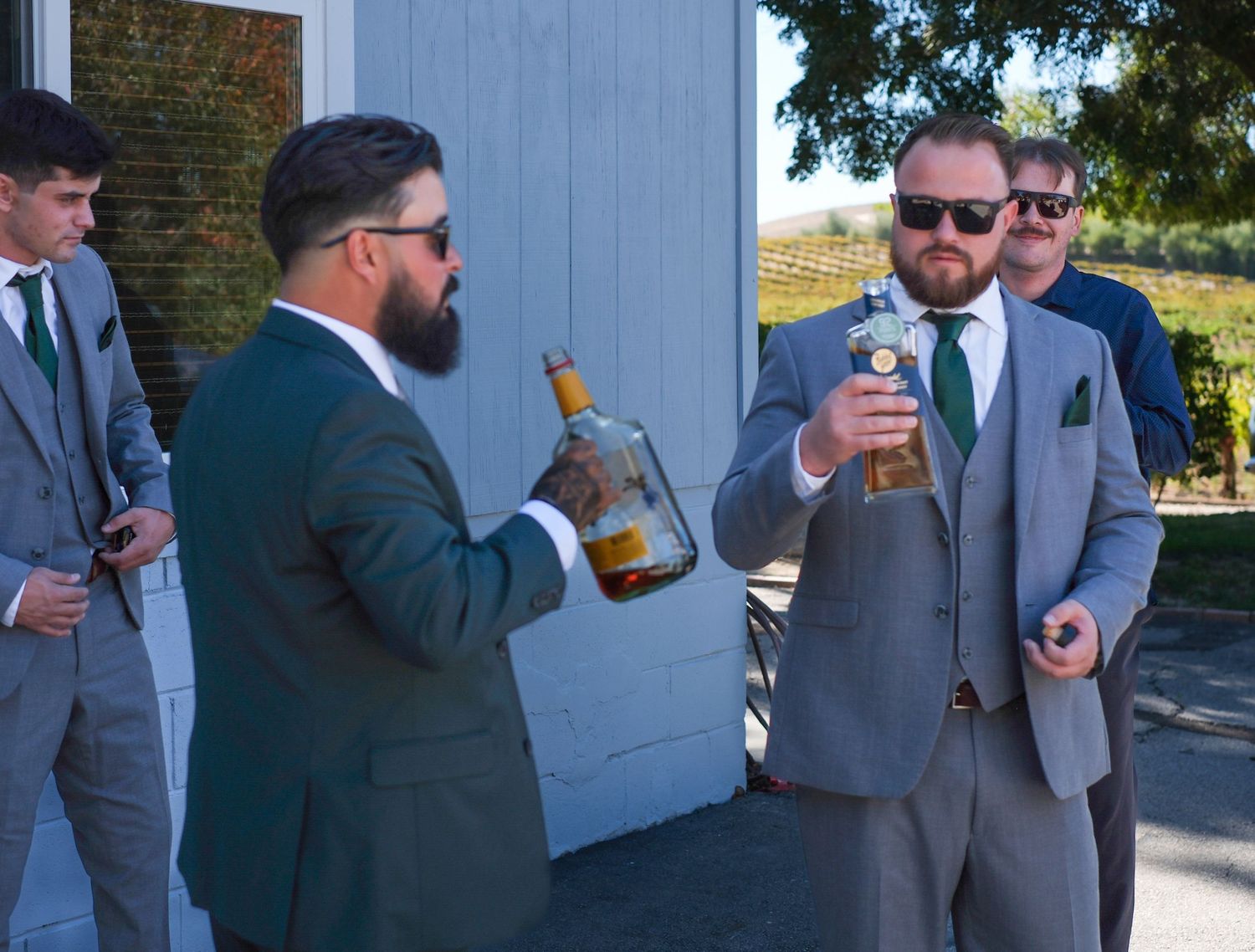 Groomsmen in gray suits drinking beers together outdoors before a wedding celebration.