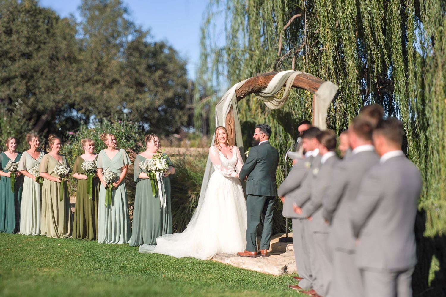 Wedding party poses under weeping willow trees on a sunny day for outdoor group photos.