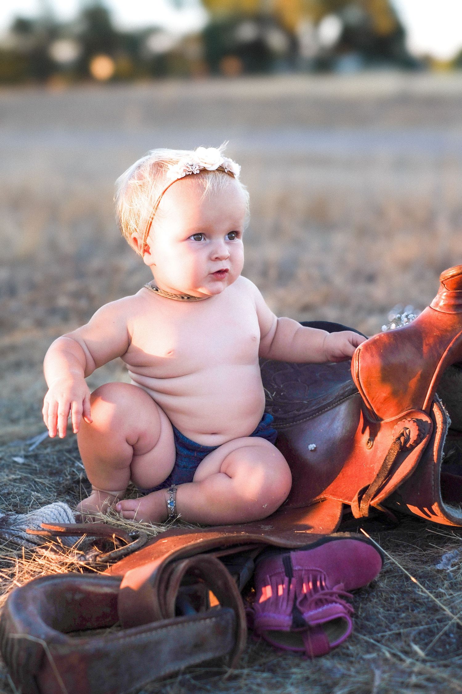 Baby wearing a headband sits on ground next to brown leather saddle and shoes in golden afternoon light.