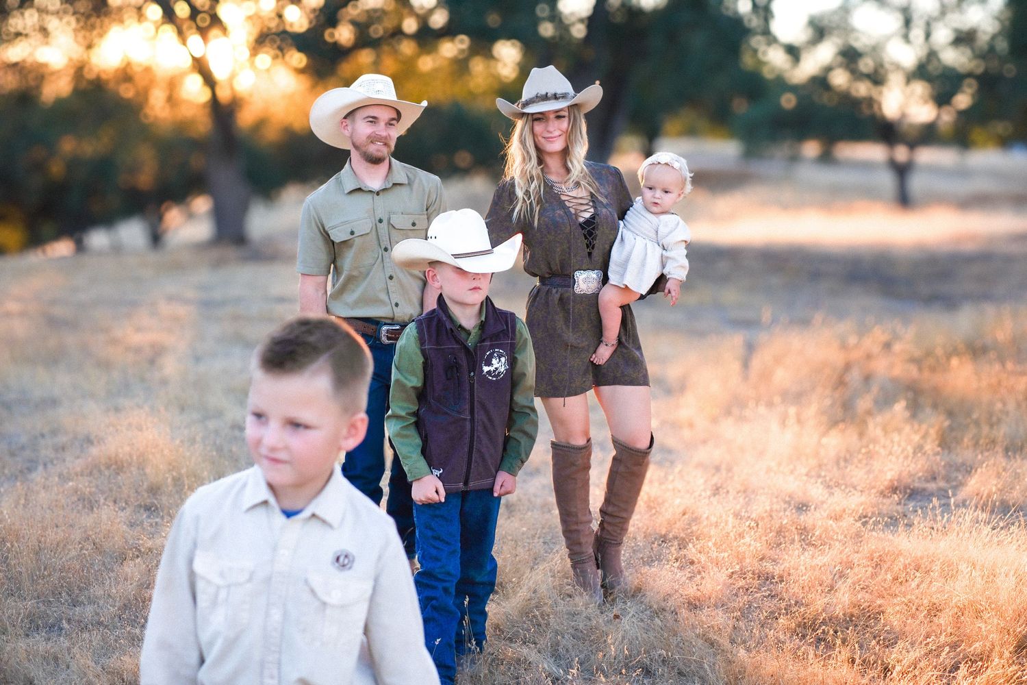 A family wearing western attire walks together through a sun-lit golden field at sunset.