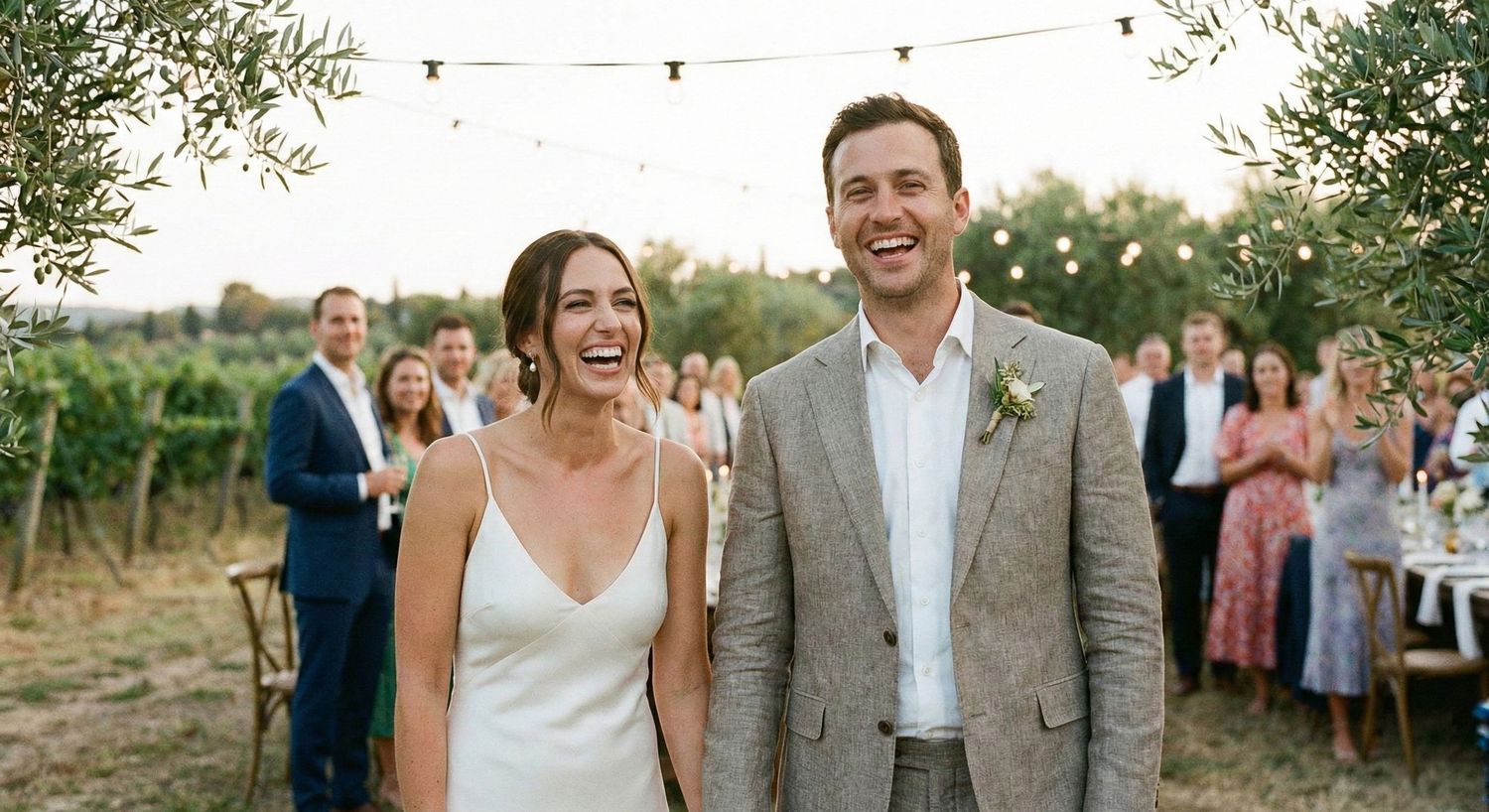Happy couple celebrating their wedding at an outdoor vineyard reception with string lights and guests in background.