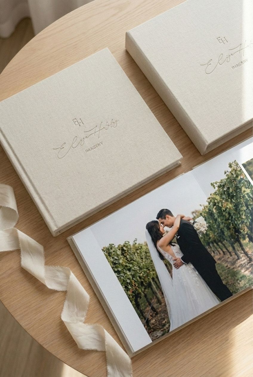 A white wedding photo album with a matching box lies open on a light wooden surface showing a romantic outdoor portrait.